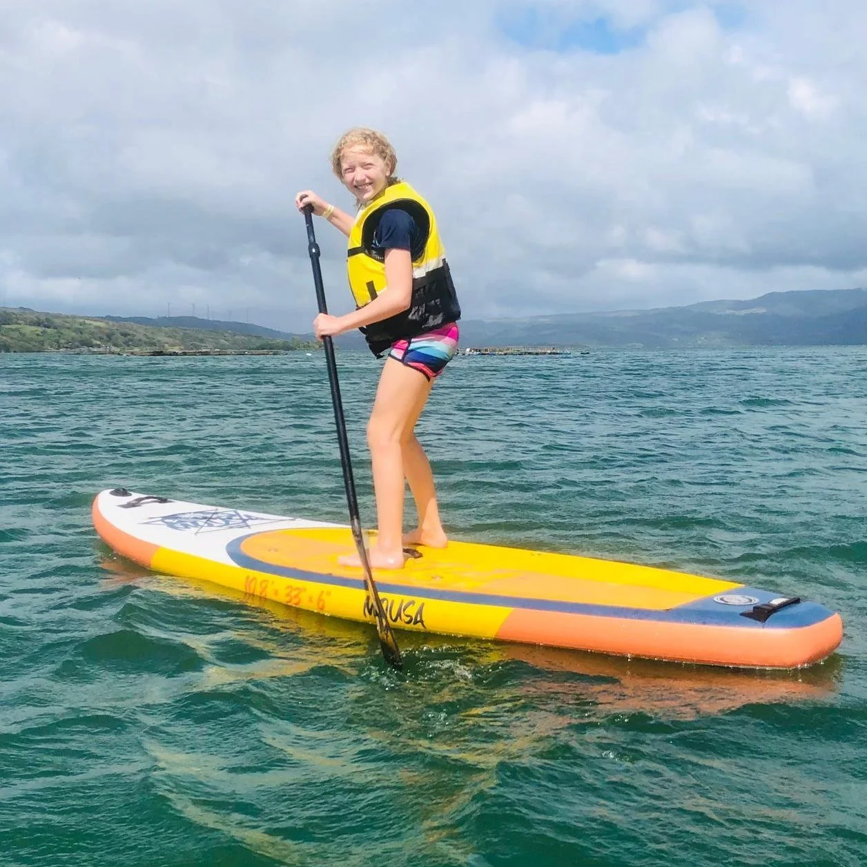 An elementary student paddleboarding on a body of water, smiling, wearing a life jacket and colorful shorts, with mountains in the background during a school field trip in Costa Rica