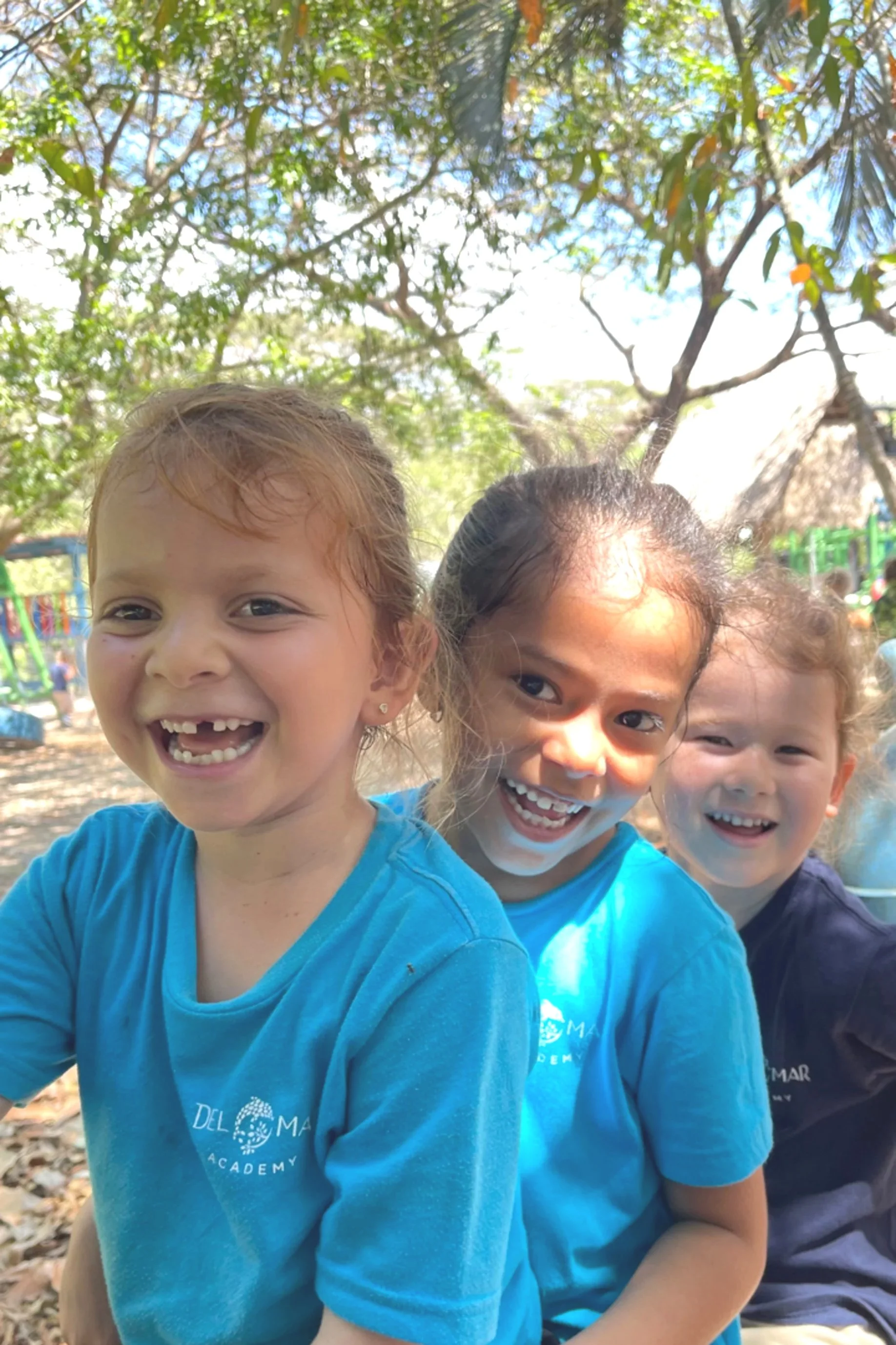 Three young girls smiling and posing for the camera outdoors, with trees and playground equipment in the background.