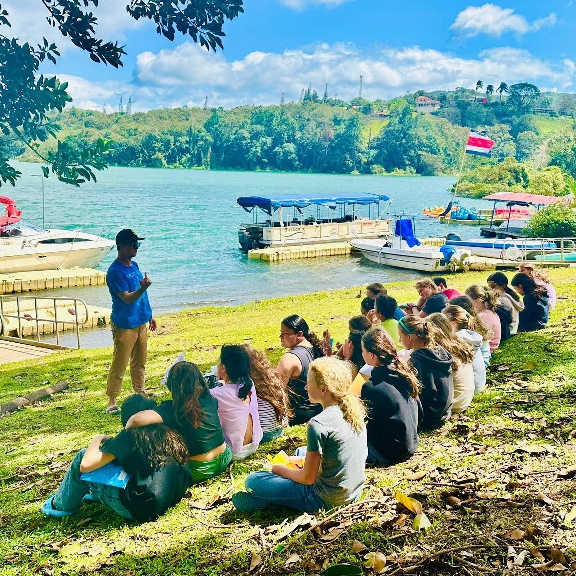 A group of middle school students sitting on the grass by a lake, listening to an instructor. There are boats docked on the lake, with a big flag flying in the background under a partly cloudy sky in Costa Rica