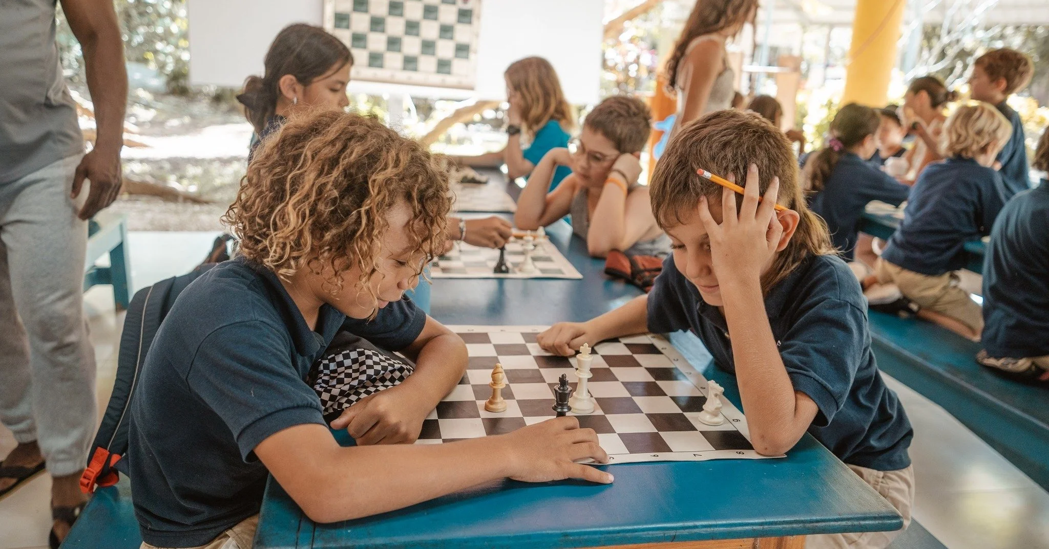 Children playing chess at a long table in a brightly lit indoor space, with some children watching and others focused on their game.