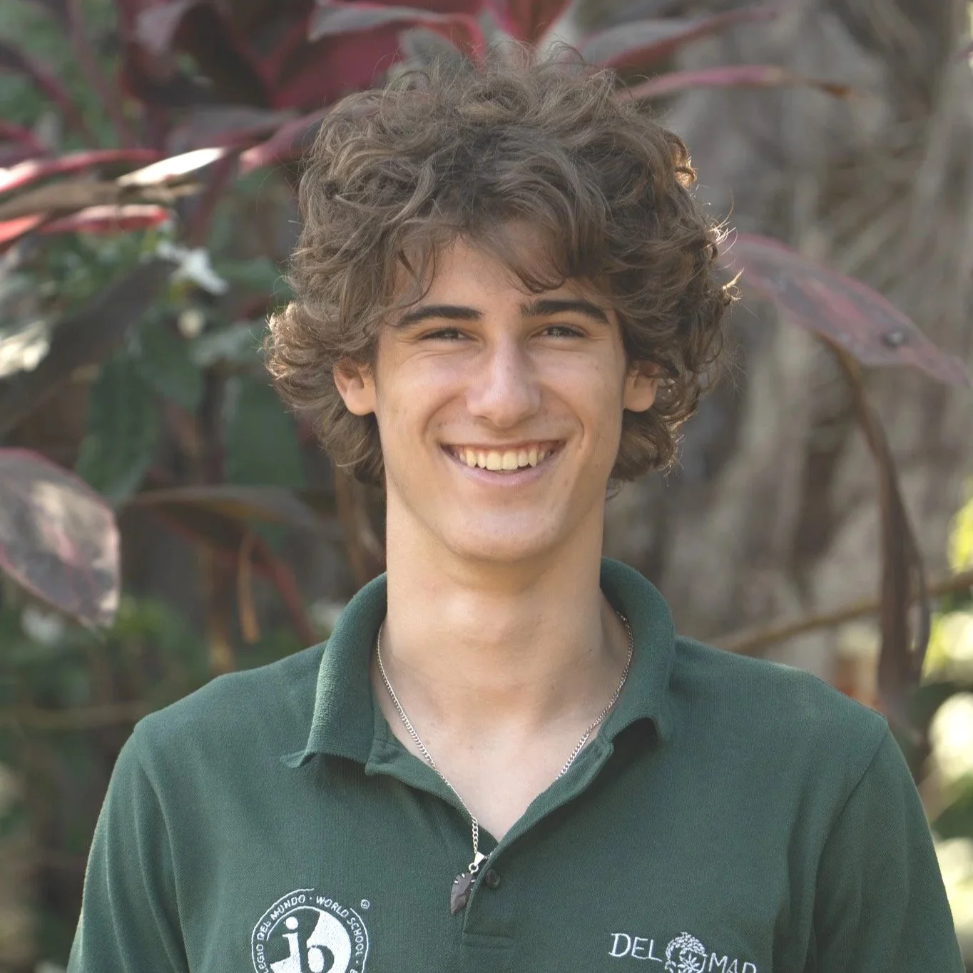 A young man with curly brown hair smiling outdoors with plants in the background, wearing a green polo shirt with logos.
