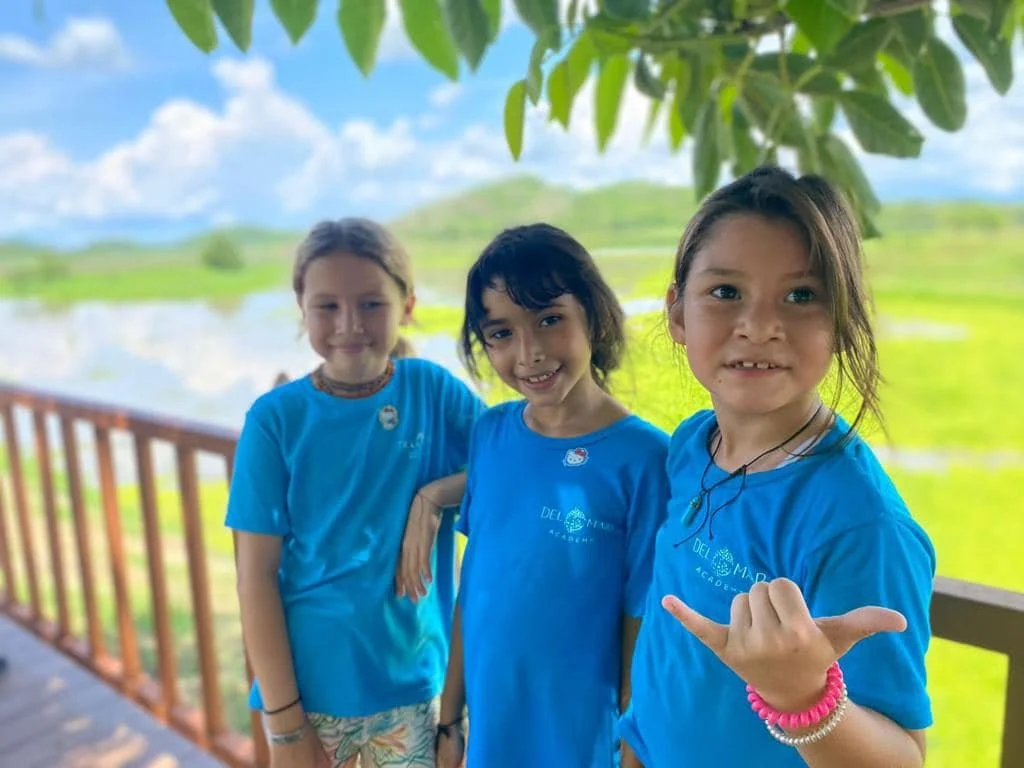 Three kindergarten students standing outdoors on a deck with a railing, wearing matching blue t-shirts, lush green landscape, water, and hills in the background, under a partly cloudy sky in Costa Rica during a school field trip