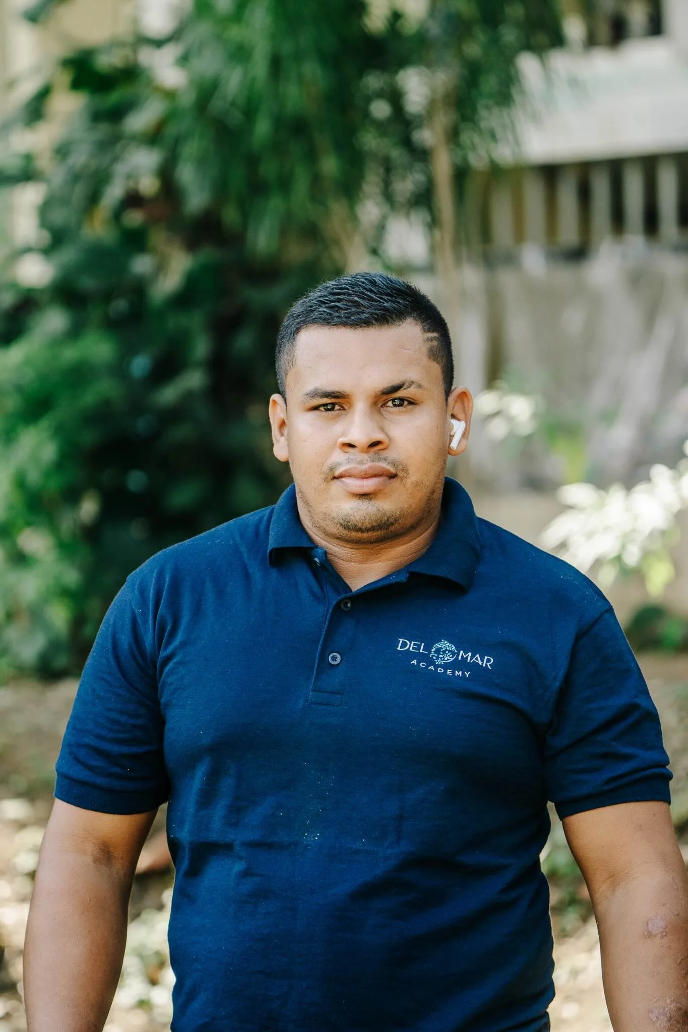 A young man with short black hair, wearing a navy blue polo shirt with 'Del Mar Academy' embroidered on it, standing outdoors with green foliage and a blurred background.