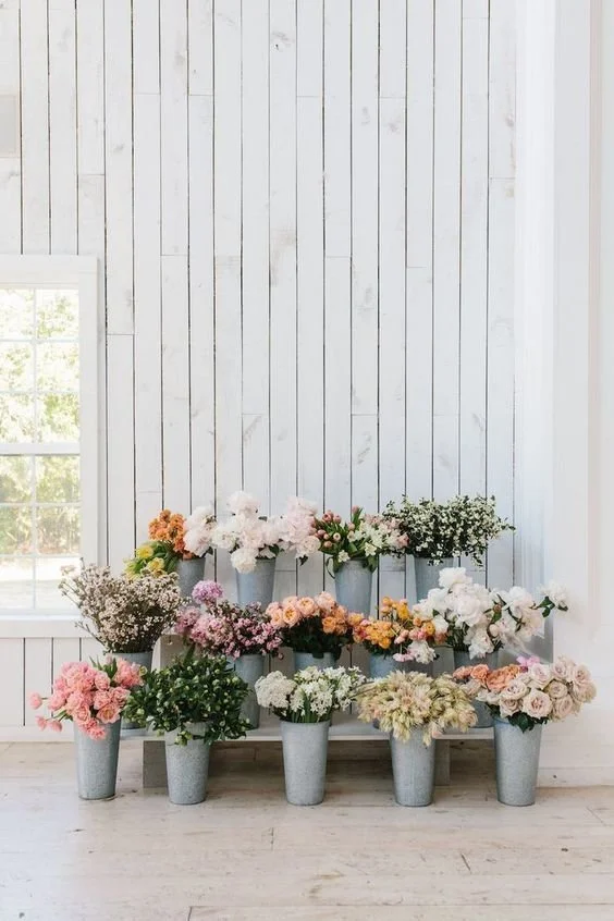 Multiple vases with colorful flowers are arranged on a wooden stand against a white wooden wall.