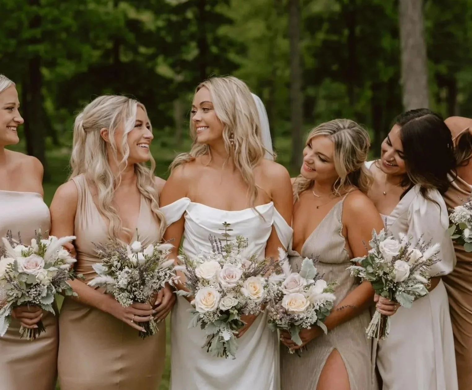 A group of women dressed in beige and white dresses holding bouquets, standing outdoors in a wooded area, smiling and interacting with each other.