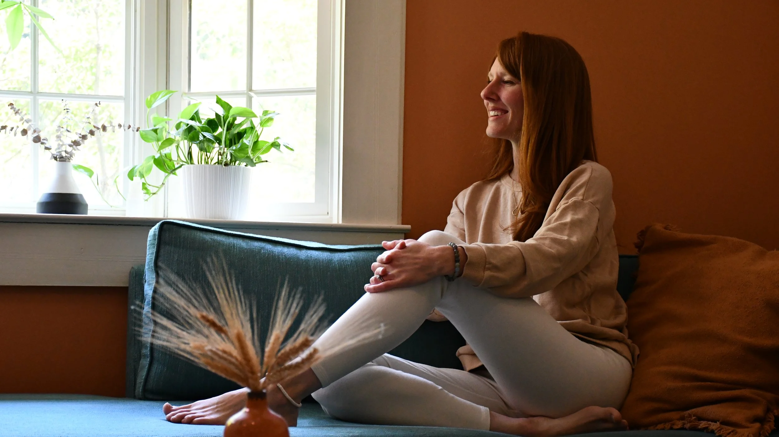 A woman with red hair sitting on a couch near a window, smiling with her eyes closed, with plants on the windowsill and a dried plant in a decorative vase in the foreground.