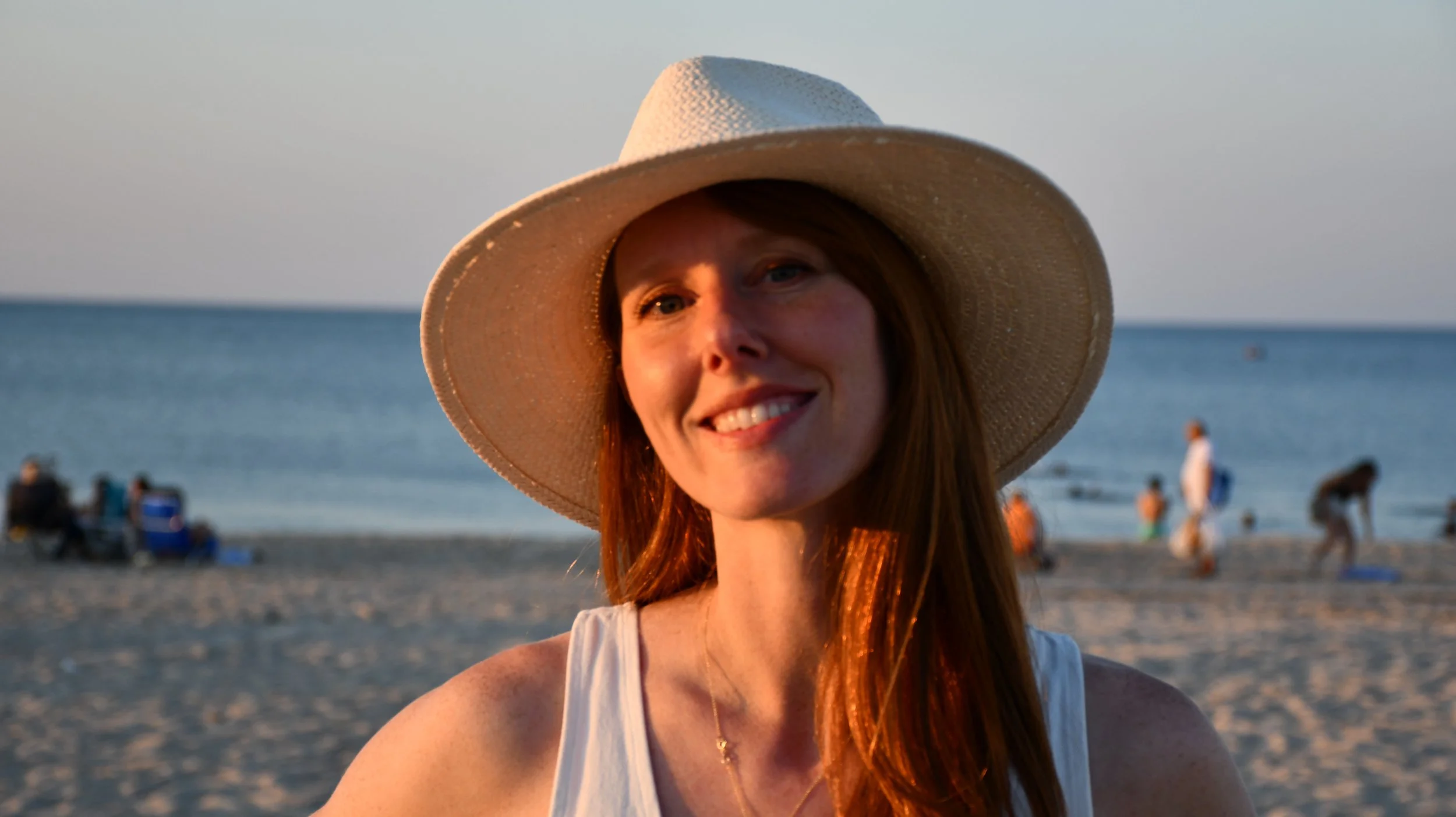A woman with red hair wearing a wide-brimmed hat and white top, smiling at the beach during sunset.