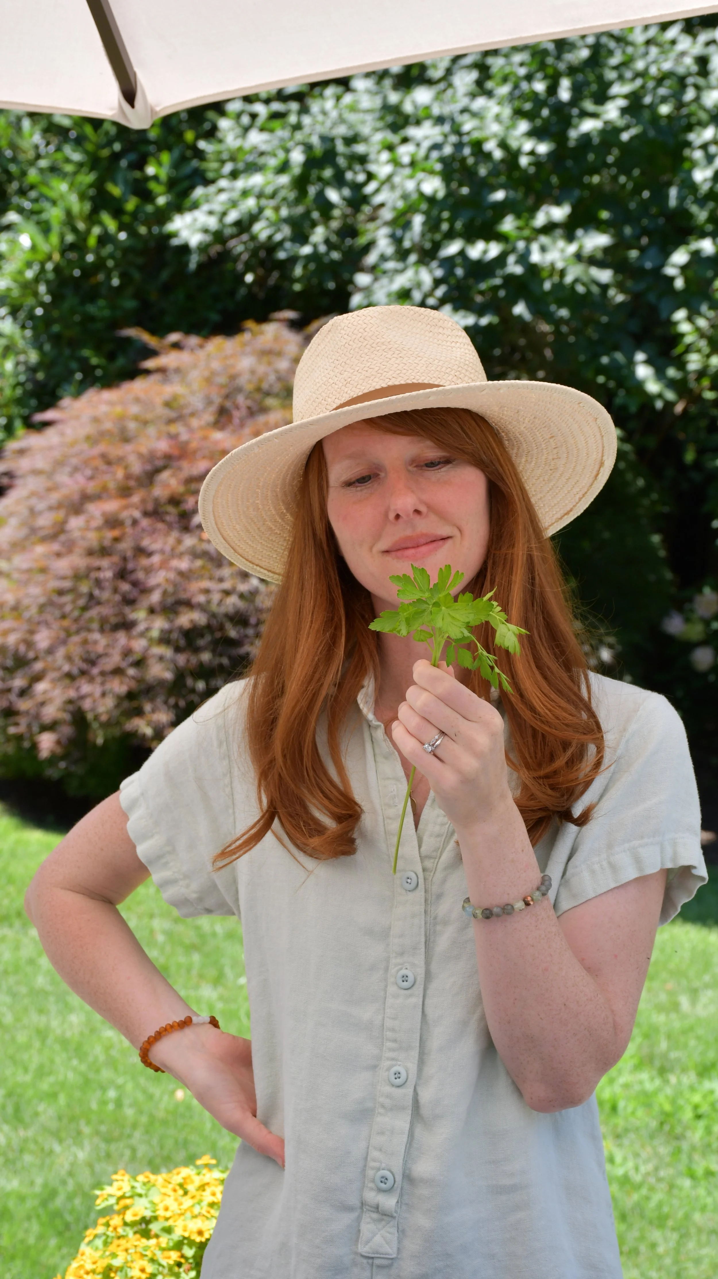A woman with long red hair, wearing a wide-brimmed straw hat and a light beige button-up shirt, is holding a sprig of green leaves in her right hand and looking at it with a gentle expression. She is outdoors with a lush green background, including bushes and yellow flowers.
