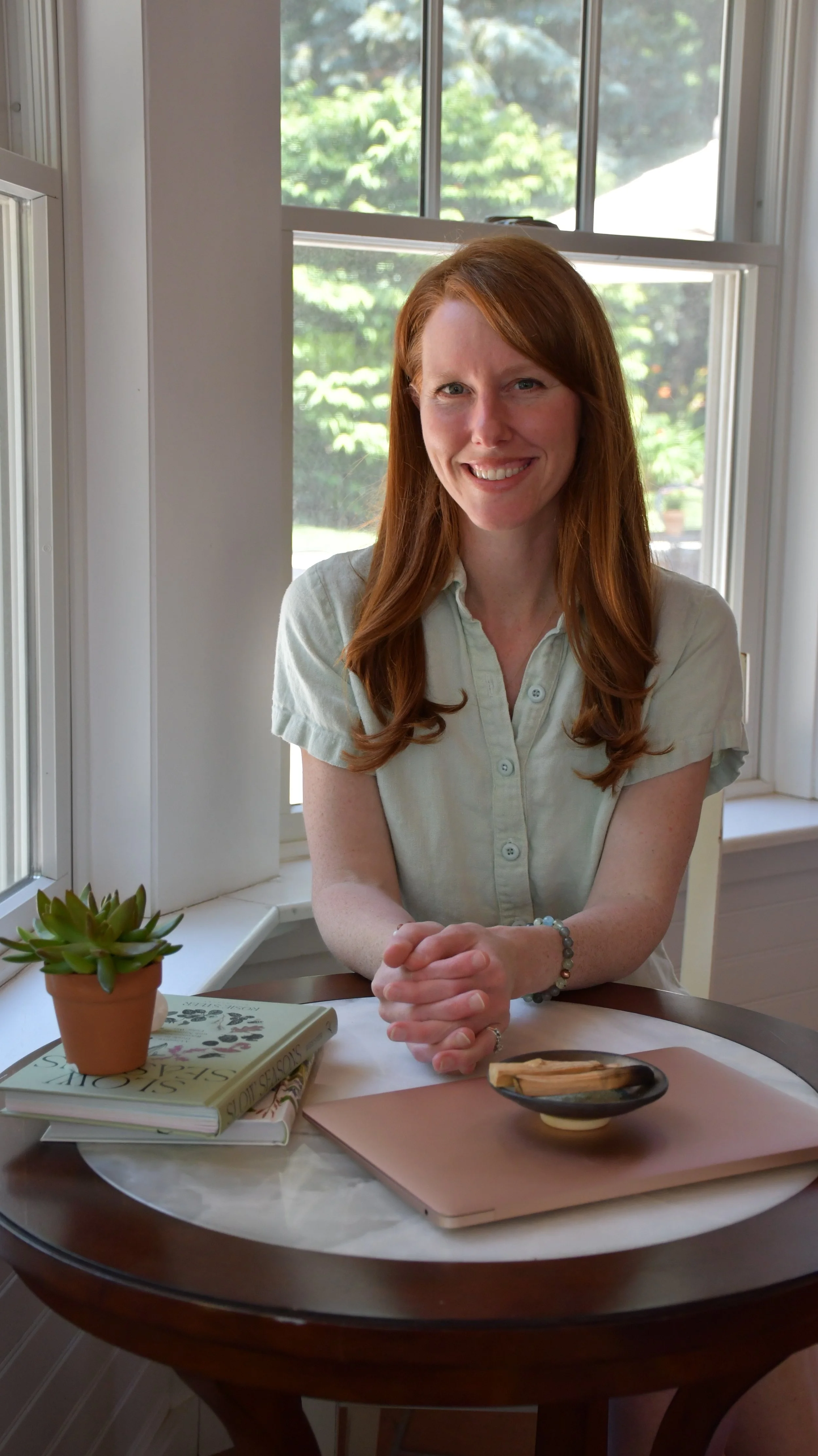 A woman with long red hair and fair skin sitting at a round dining table, smiling at the camera. She is wearing a short-sleeved, light-colored button-up shirt and a bracelet on her wrist. The table has a closed laptop, a small potted succulent, a stack of books, and a decorative bowl. Behind her, a window shows a sunny outdoor scene with trees.