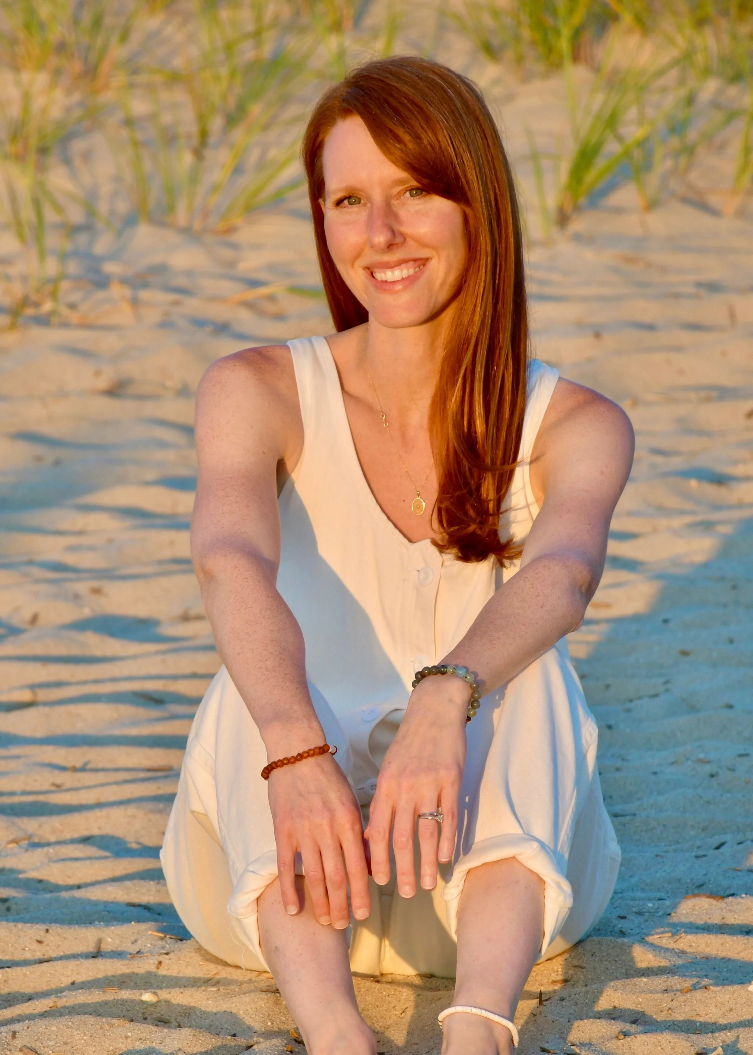 A woman with long red hair smiling while sitting on a sandy beach with grass in the background, wearing a white sleeveless dress and jewelry.