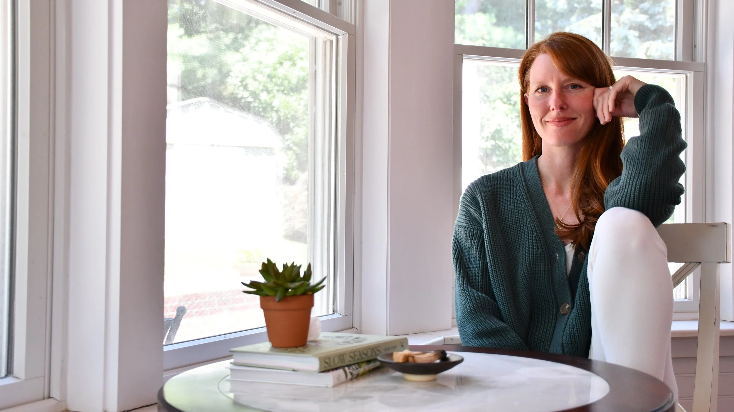 A woman with red hair sitting at a dining table near a window, smiling and resting her head on her hand.