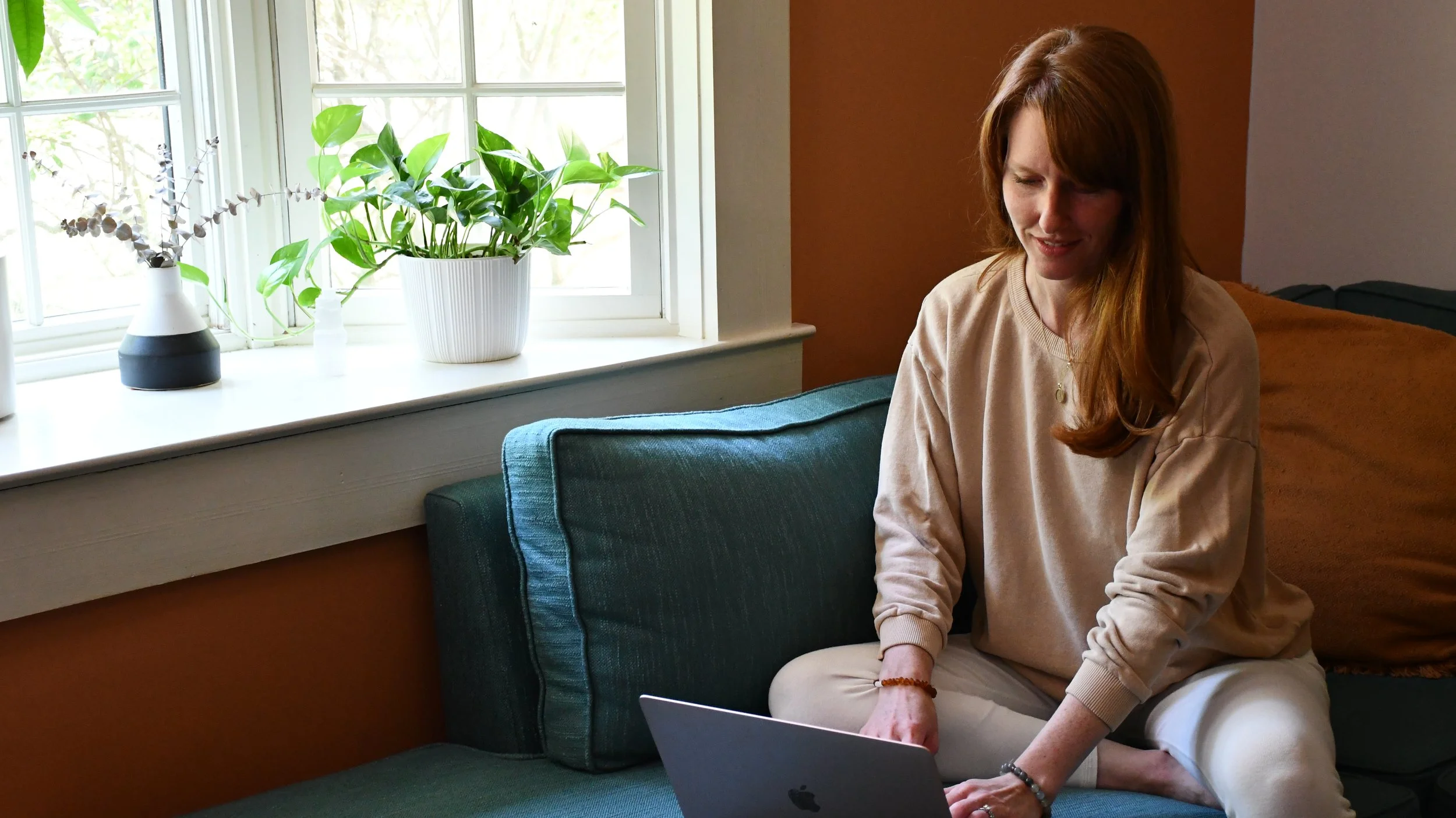 A woman with red hair sitting on a blue-green couch by a windowsill with green plants and a white and black vase, working on a silver laptop in a cozy room.