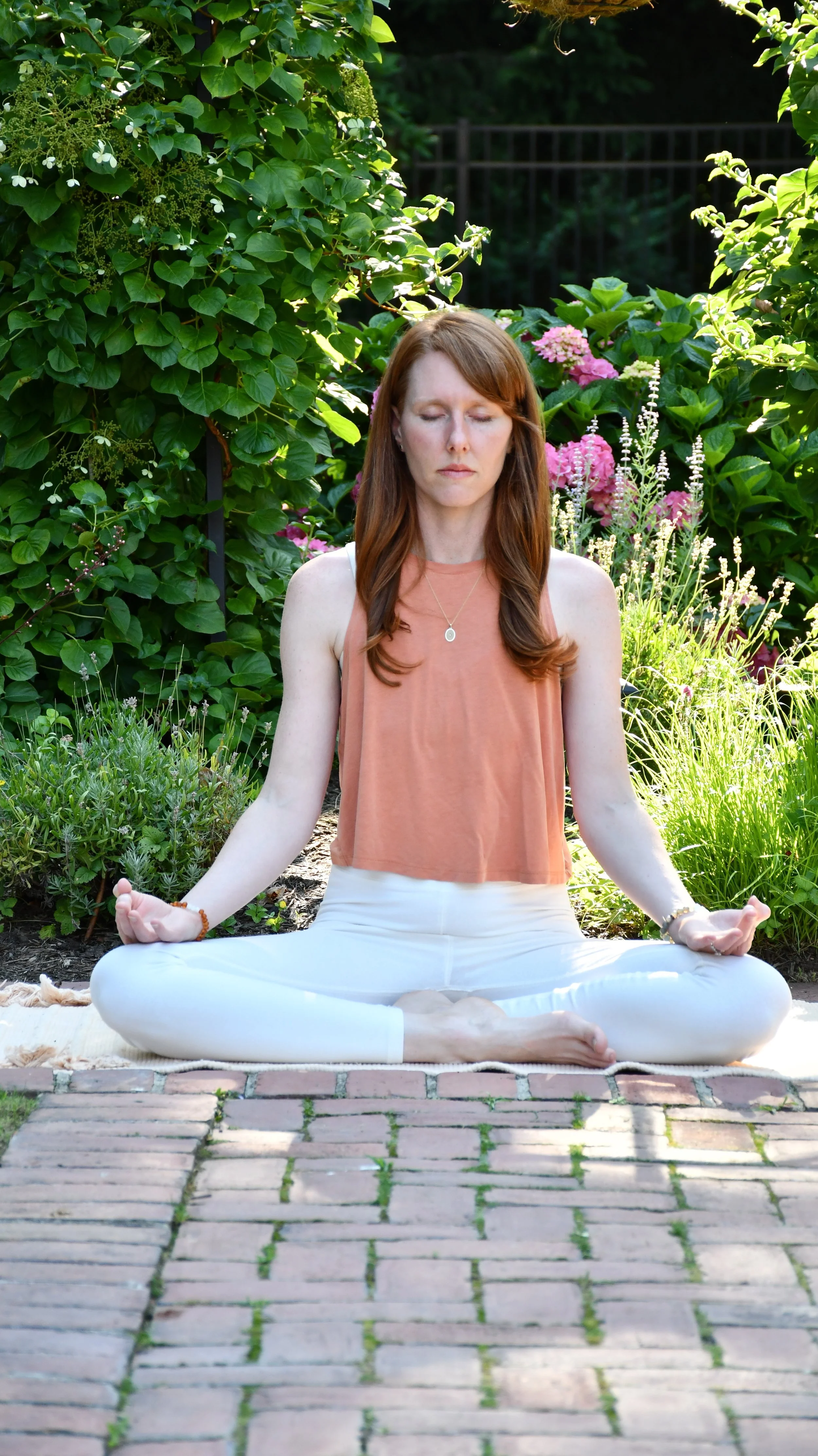 A woman practicing yoga outdoors on a mat, sitting cross-legged with her eyes closed, surrounded by lush green plants and flowers in a garden.