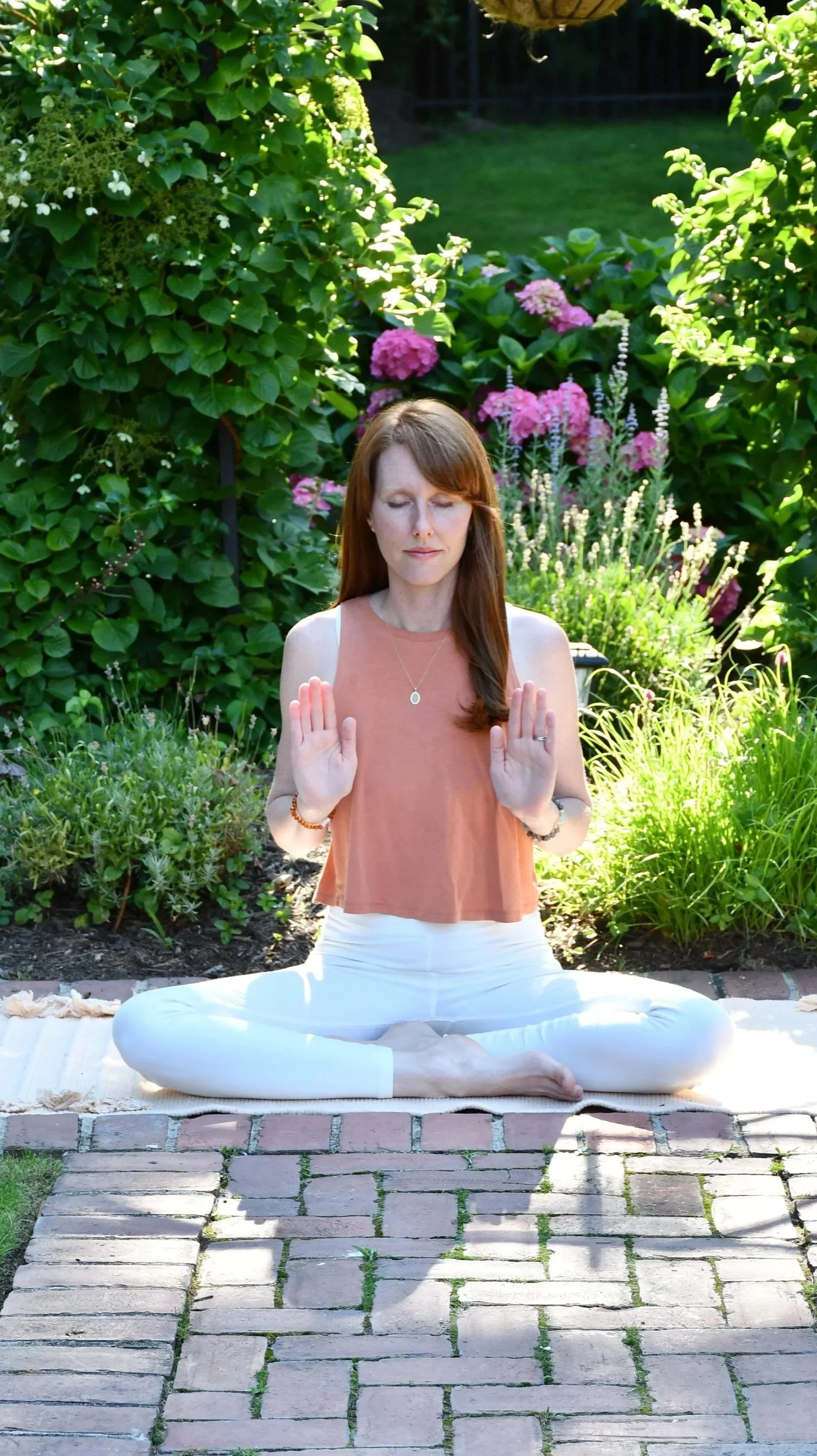 A woman practicing yoga outdoors. She is sitting cross-legged on a yoga mat on a brick patio, with her eyes closed and hands in a prayer position. She is surrounded by lush green plants and pink flowers.