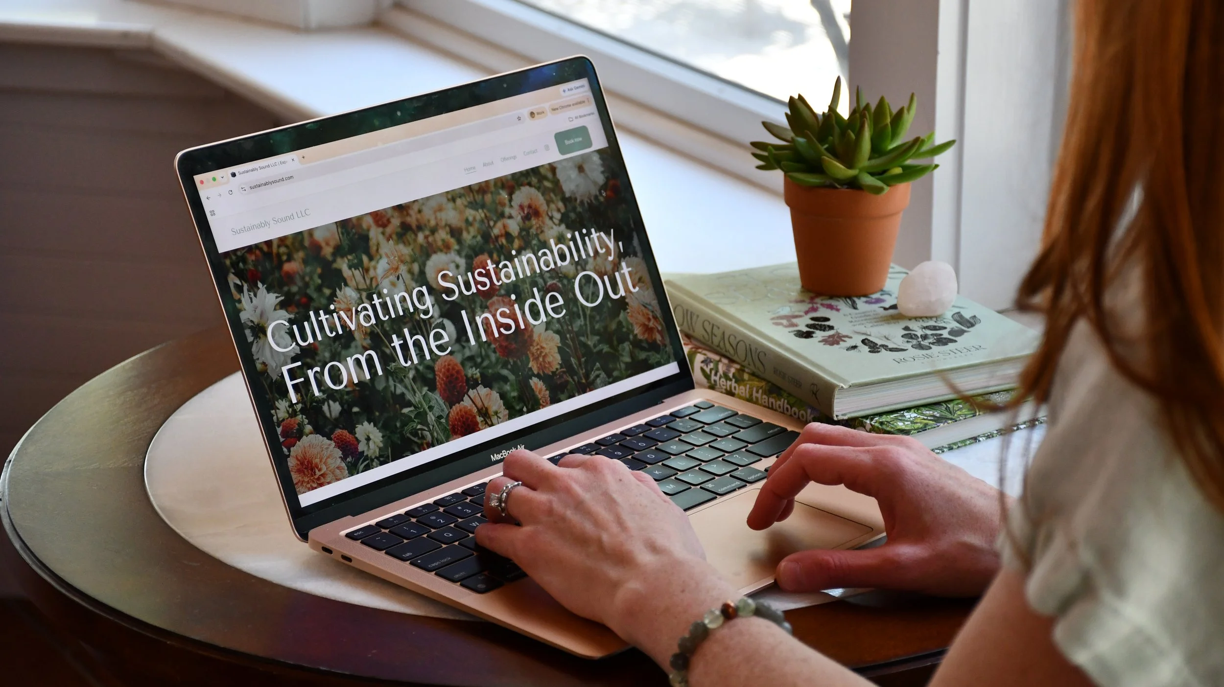 A person using a laptop on a round table, viewing a website titled 'Sustainably Sound LLC' with the slogan 'Cultivating Sustainability From the Inside Out.' There are books about herbalism and a potted succulent plant on the table, with a window in the background.