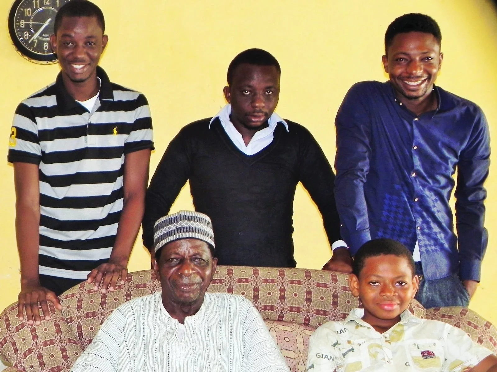 Group photo of six people, five standing behind a sofa with one seated, in front of a yellow wall with a clock showing 9:40.