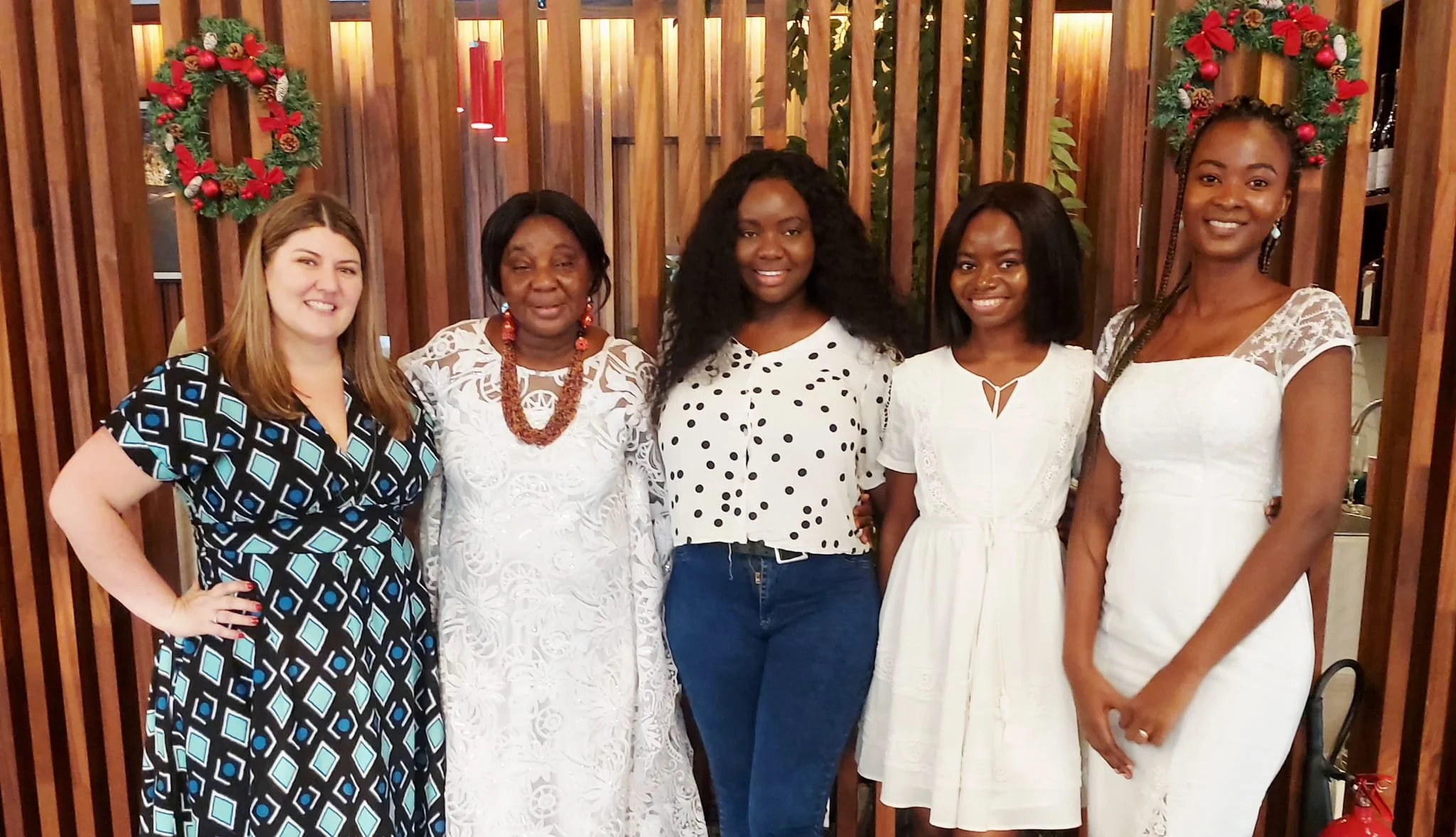 Five women standing together in a restaurant, dressed in white and blue, with Christmas wreath decorations in the background.