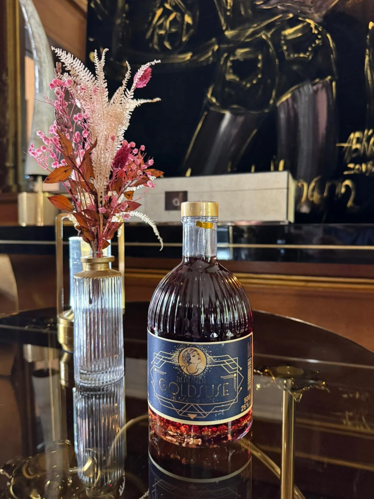 Eine Flasche mit dunkler Flüssigkeit und ein Blumenstrauß in einer Schmalen Glasvase auf einem schwarzen Tisch.