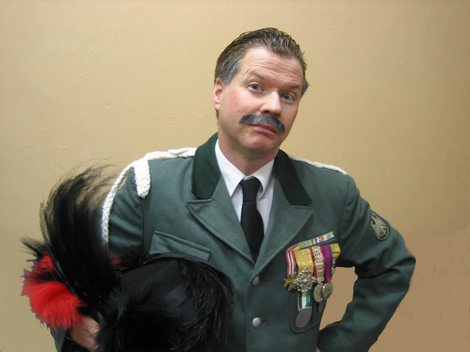 Ian Prosser as Albert Bertorelli dressed in a decorated green military uniform with medals and ribbons, holding a black and red feathered hat, standing against a plain beige wall.