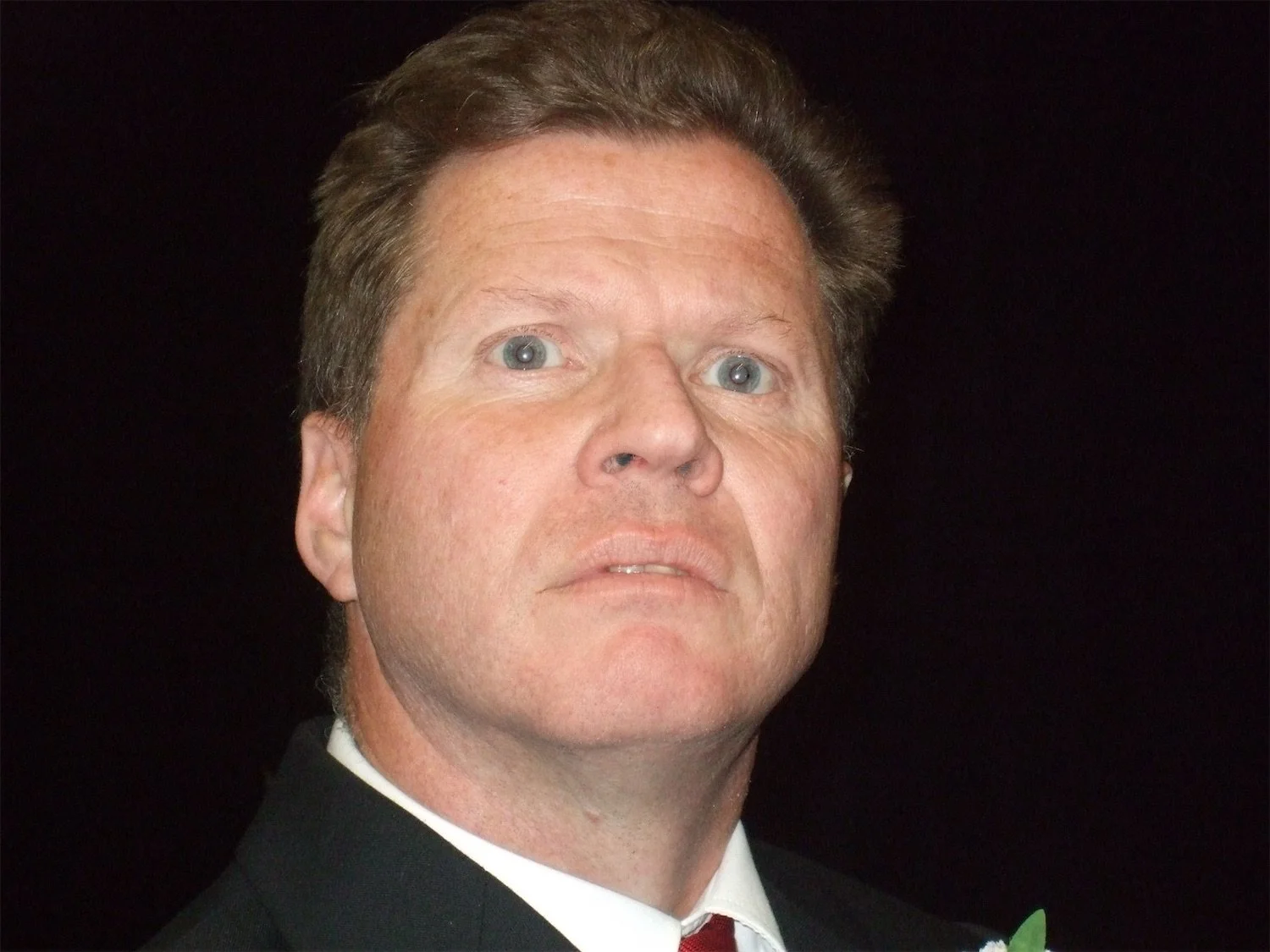 Close-up photo of actor Ian Prosser in character with light skin, blue eyes, brown hair, wearing a tuxedo and red tie, against a black background.