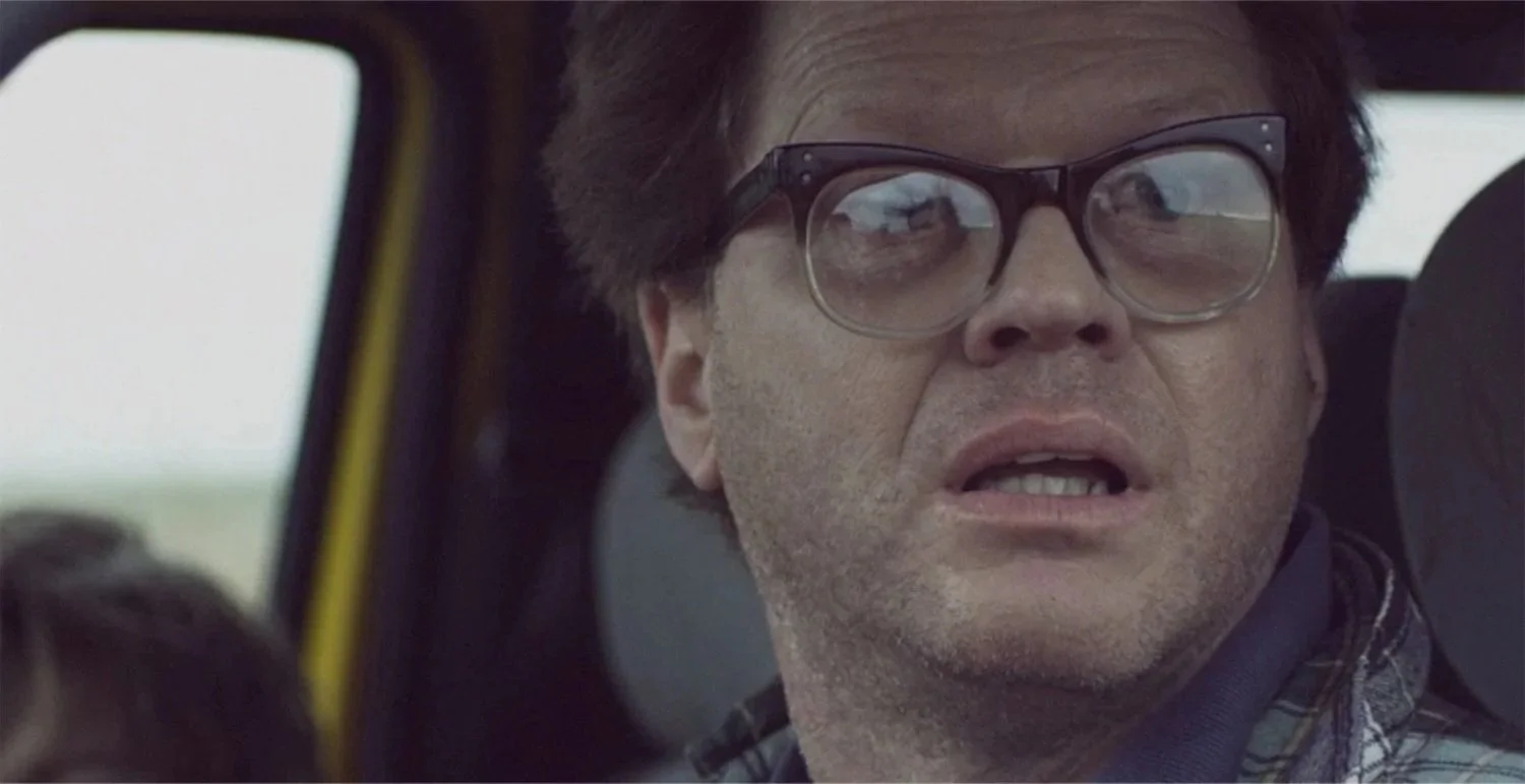 Actor Ian Prosser in character with glasses and brown hair looking out the window of a vehicle, with a surprised or concerned expression.
