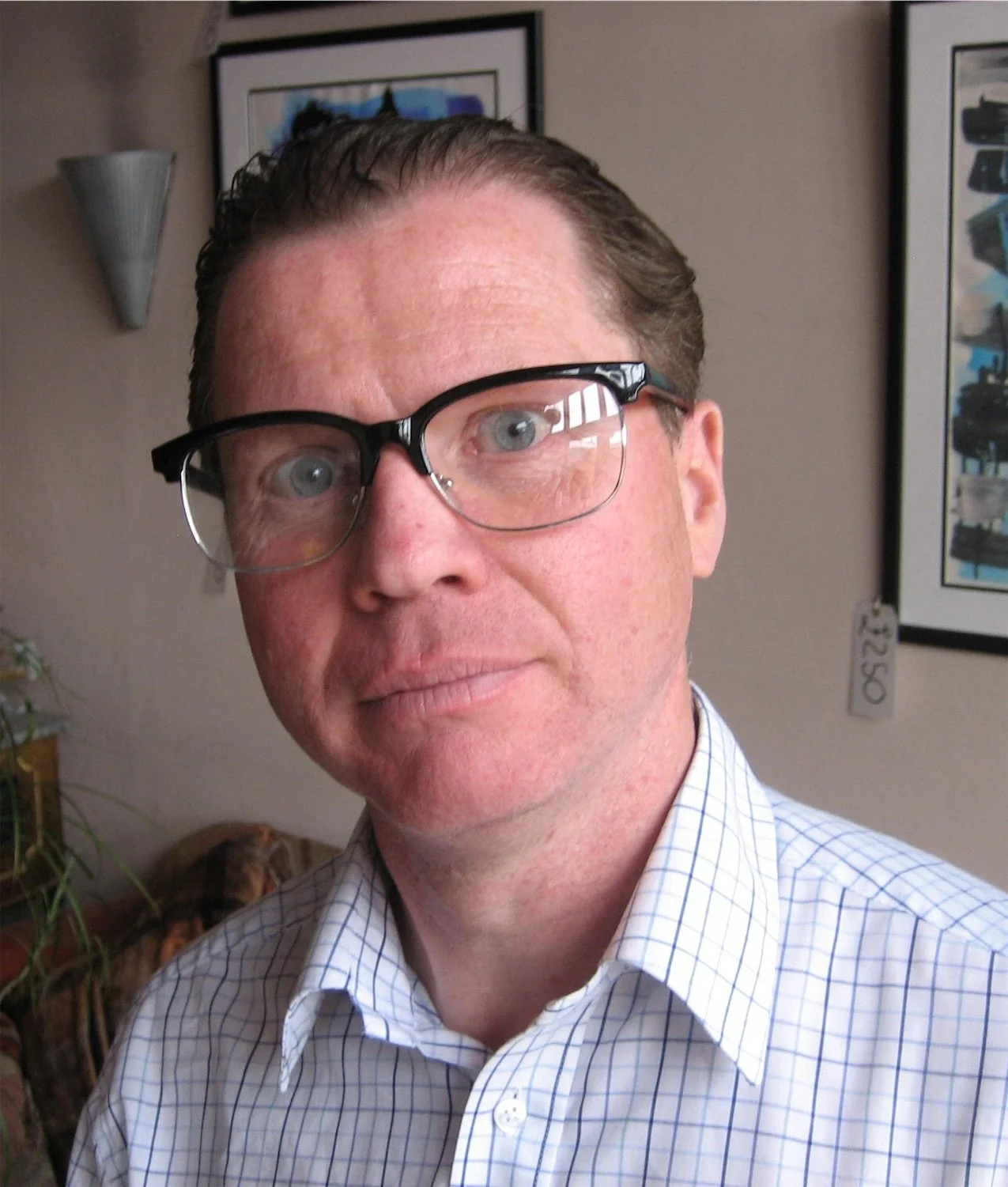 Close-up of actor Ian Prosser in character with glasses, wearing a checkered shirt, standing indoors with framed artwork on the wall behind him.