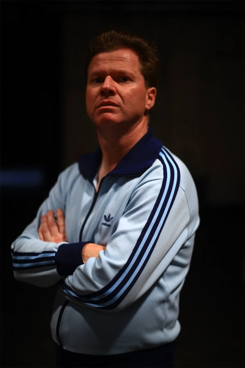 Actor Ian Prosser in character with light skin, brown hair, and a serious expression, standing with his arms crossed, wearing a white Adidas jacket with blue stripes and a blue collar, against a dark background.