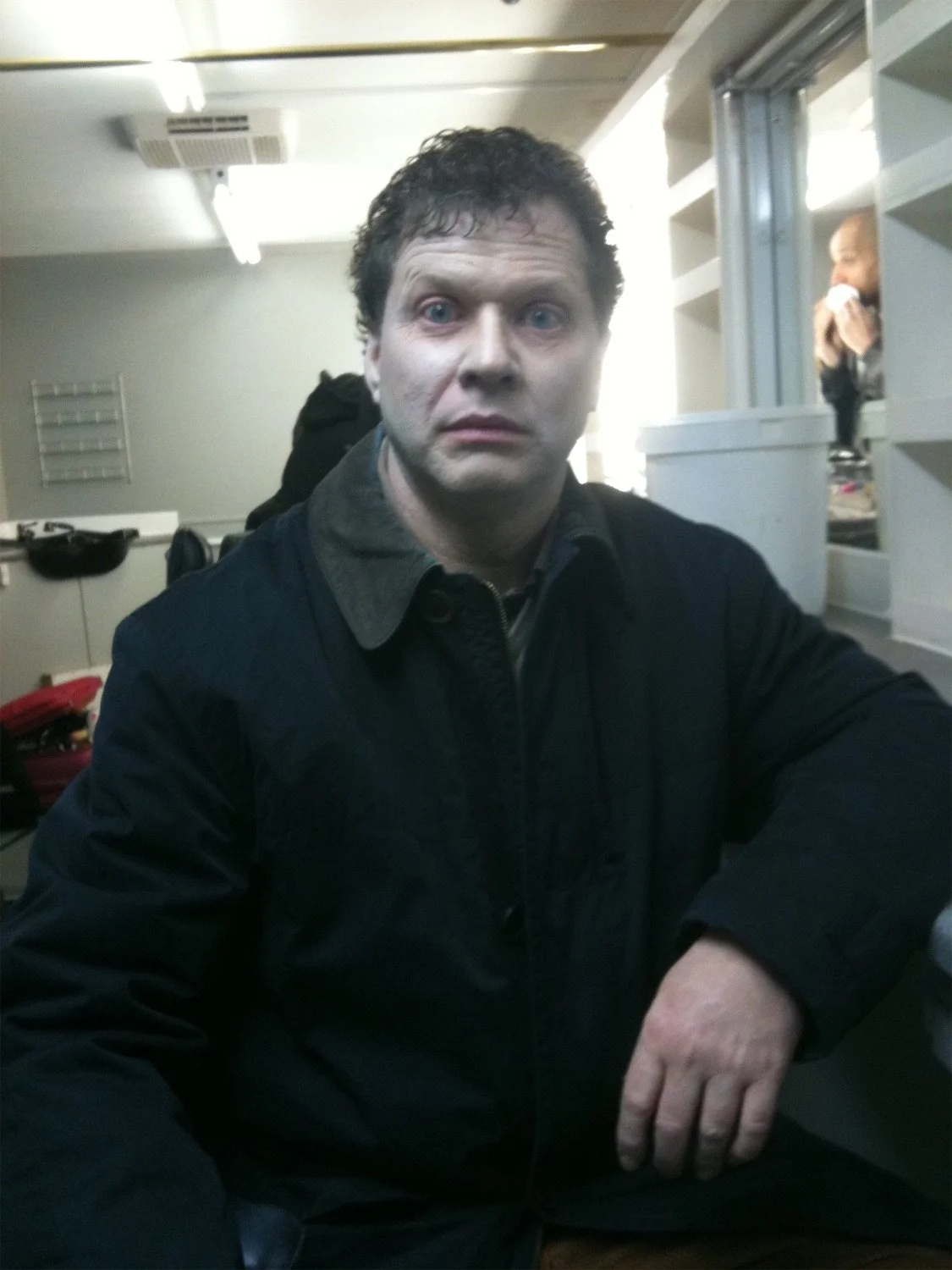 Actor Ian Prosser in character with curly hair and blue eyes wearing a dark jacket, sitting indoors in front of a white wall with bright natural light coming from the window behind him.