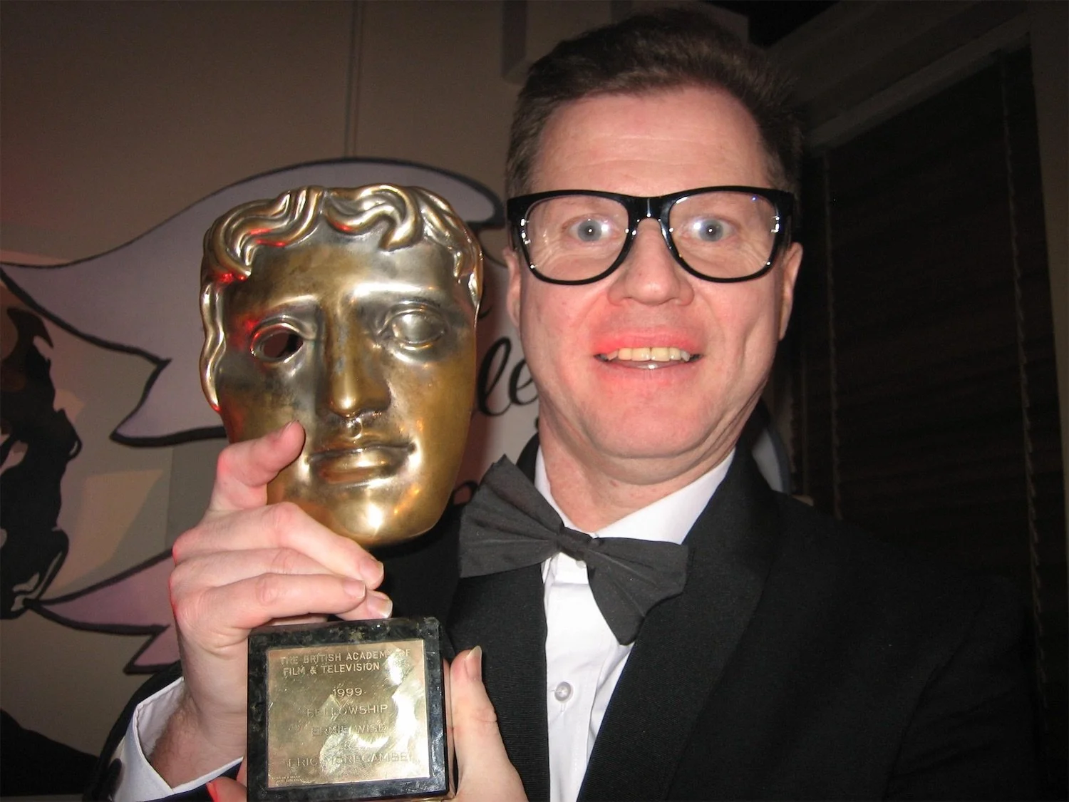 Actor Ian Prosser in character in tuxedo holding a BAFTA award smiling at the camera.