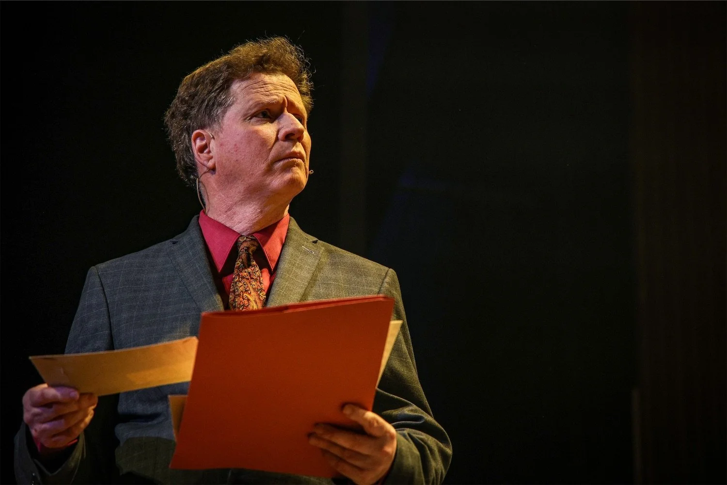 Actor Ian Prosser on stage in a grey suit holding orange and yellow folders, with a serious expression, standing against a dark background.