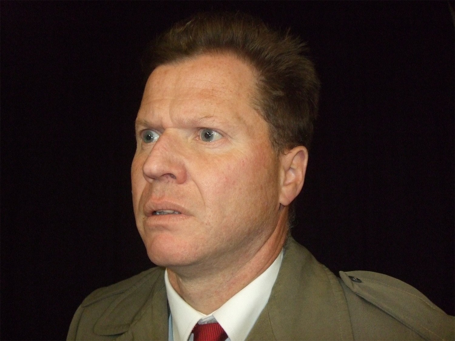 Actor Ian Prosser in character with short brown hair dressed in a mac with a red tie, looking to the left with a serious expression.
