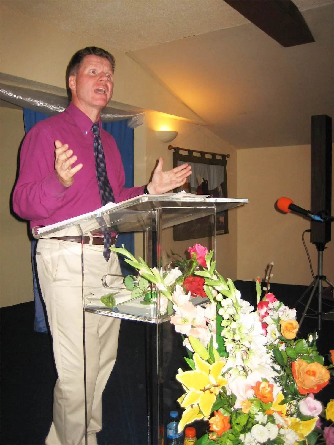 Actor Ian Prosser on stage in a performance of You Me Bum Bum Train wearing a pink shirt and tie speaking at a podium with flowers in front, in a room with a ceiling light and wall decorations.
