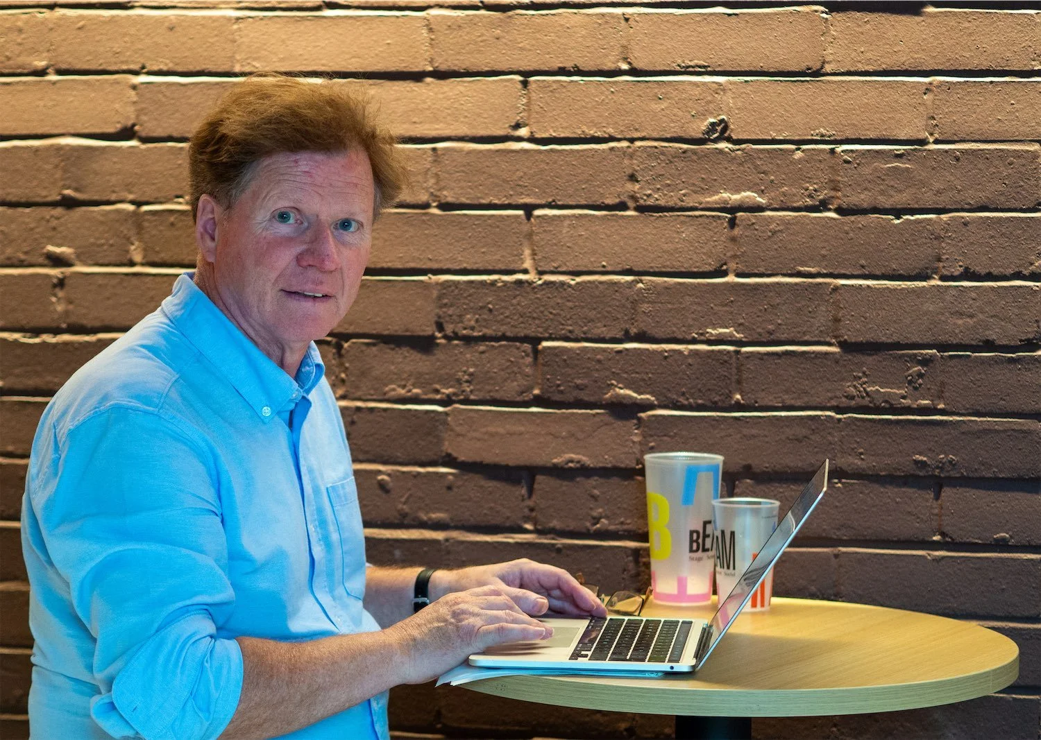 Actor Ian Prosser in a light blue shirt working on a laptop at a round wooden table in front of a brick wall, with two cups and a plate on the table.