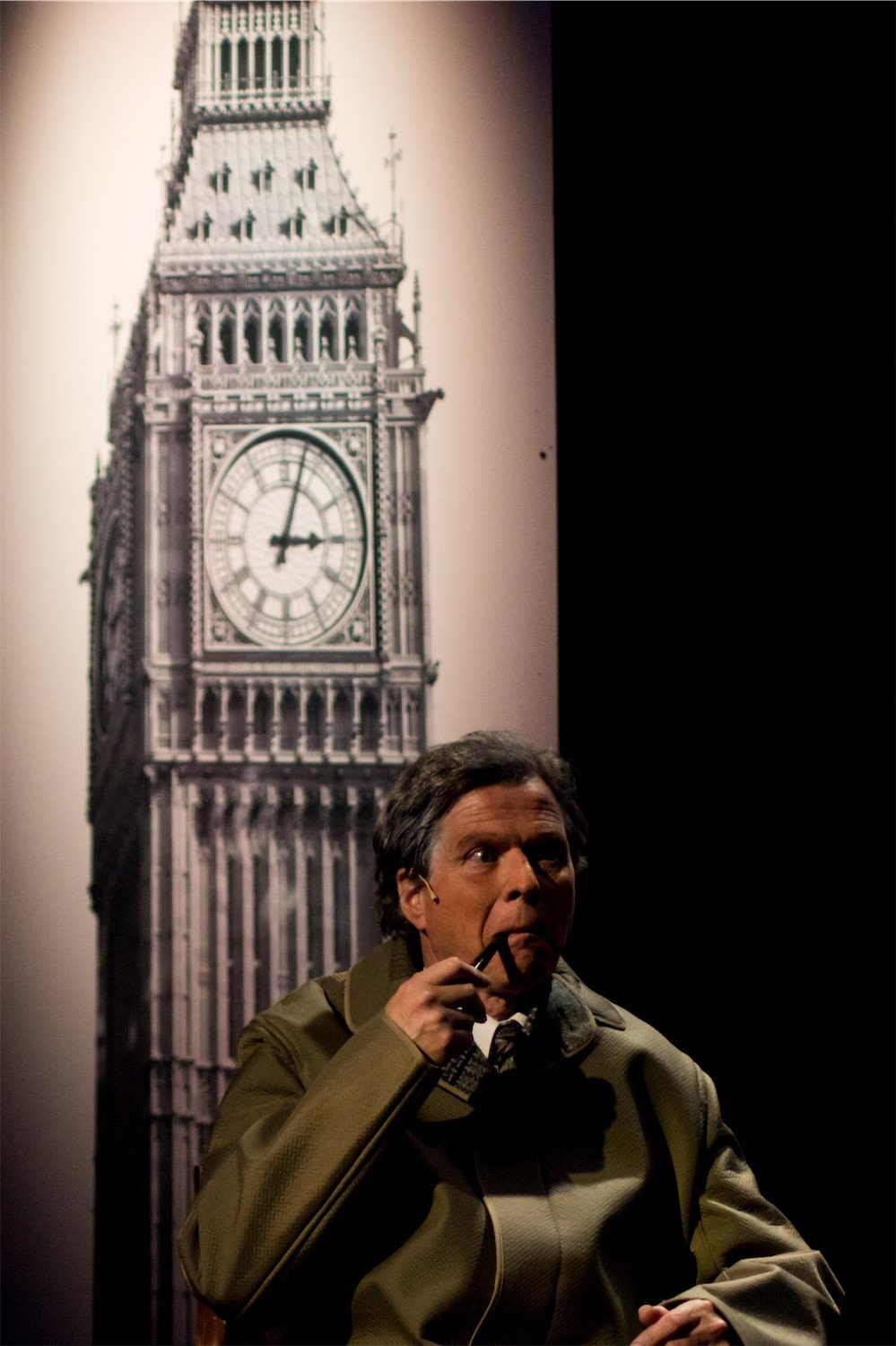 Actor Ian Prosser in character as Harold Wilson wearing a brown jacket, holding a pipe to his mouth, sitting in front of a large black-and-white image of Big Ben clock tower.