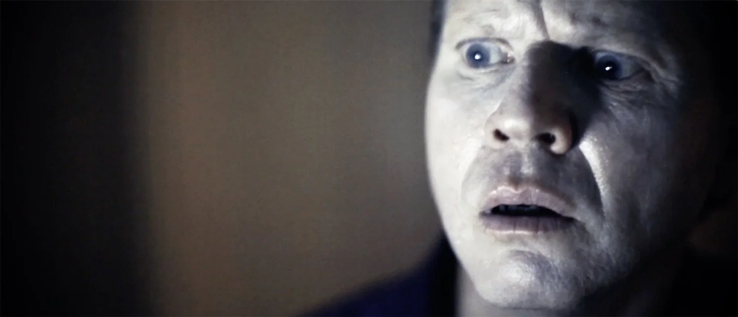 Close-up of actor Ian Prosser in character with a surprised or shocked expression, illuminated by a light source with a dark background.