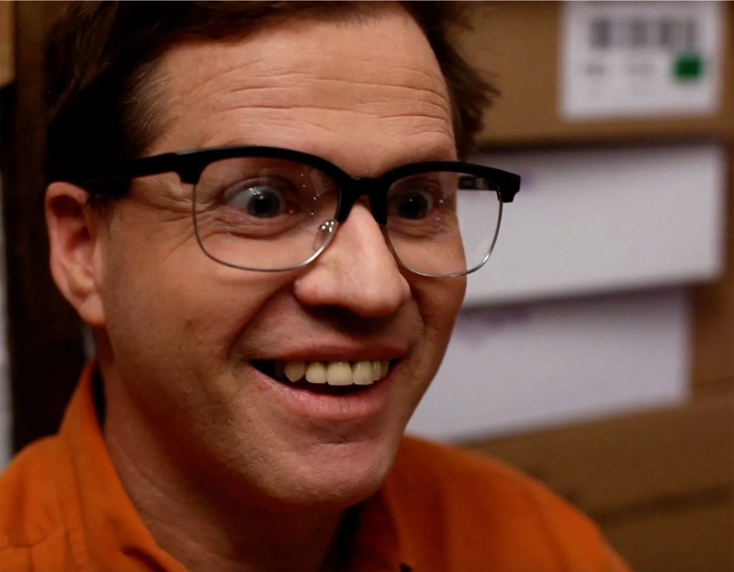 Close-up of actor Ian Prosser in character with brown hair, glasses, and an orange shirt smiling.