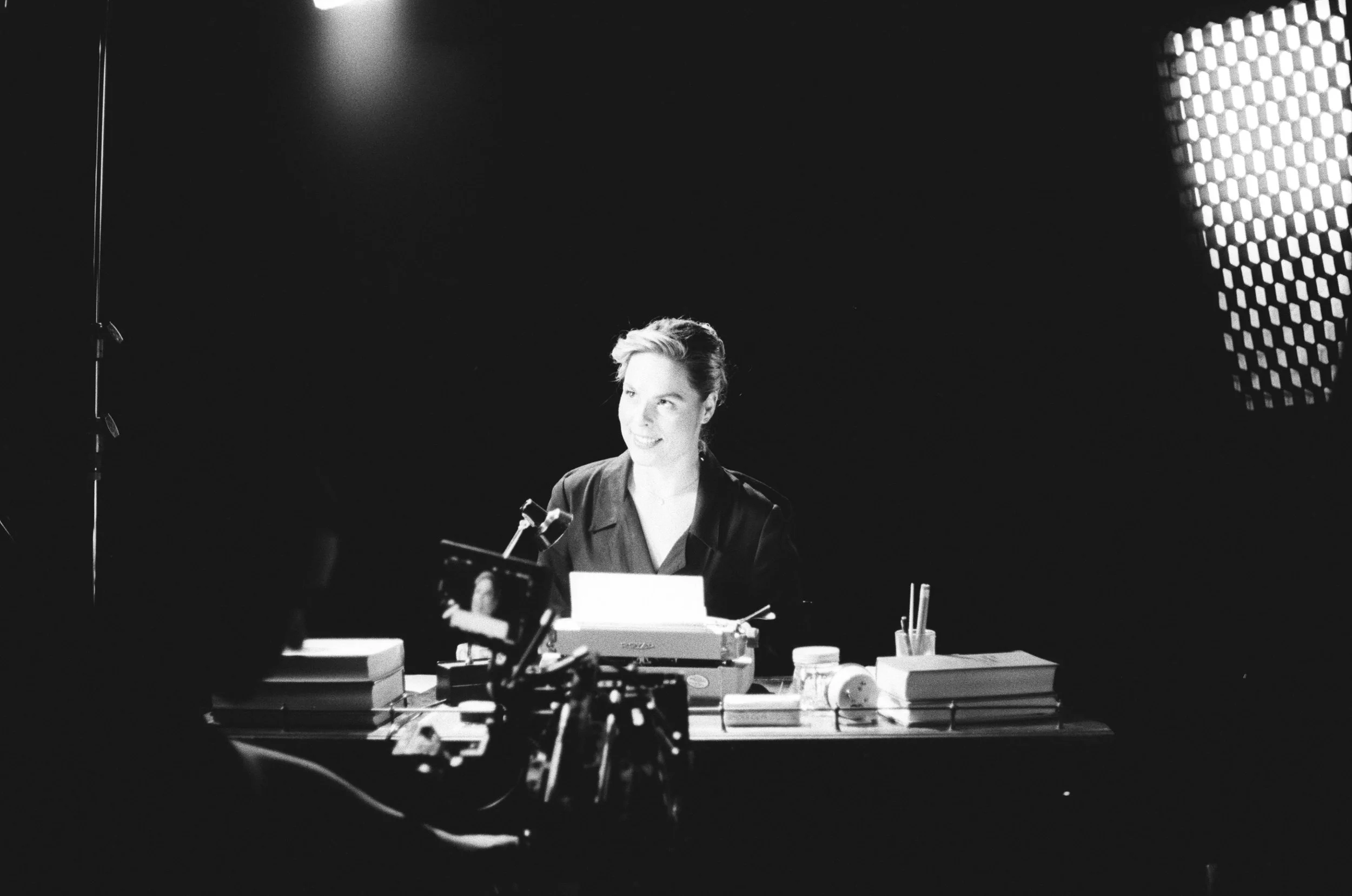 A woman with short hair sitting at a desk, smiling, in a studio setting with professional lighting and camera equipment.