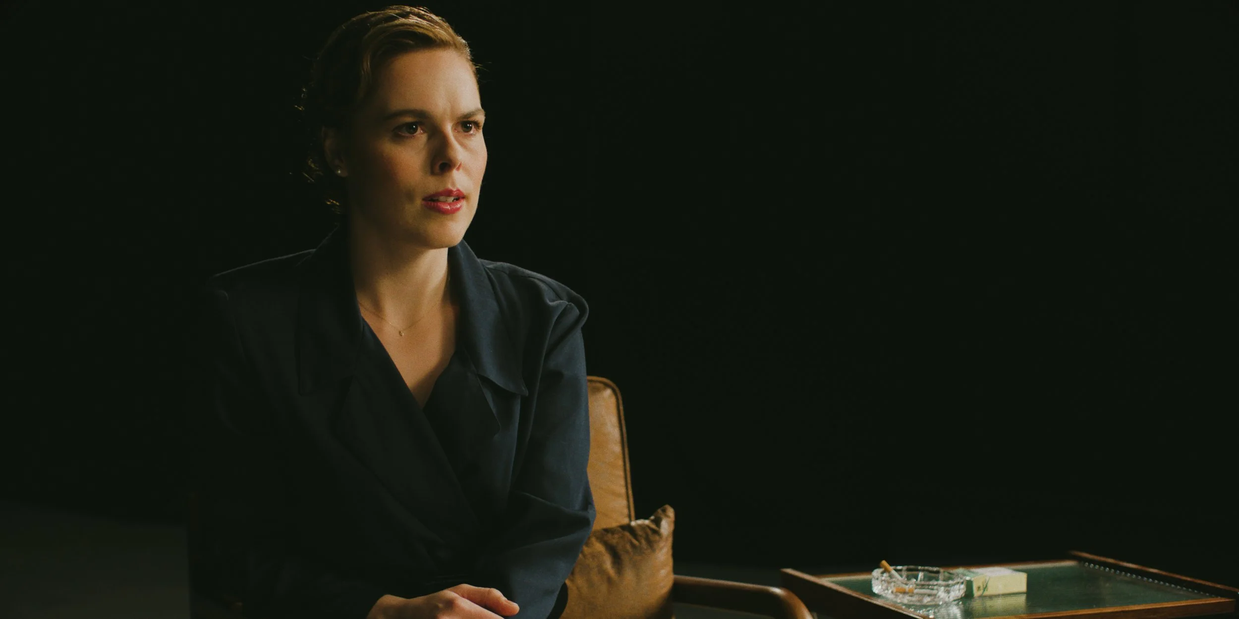 A woman with short brown hair and fair skin sitting in a dark room next to a wooden table with a pack of cigarettes, a lighter, and an ashtray.