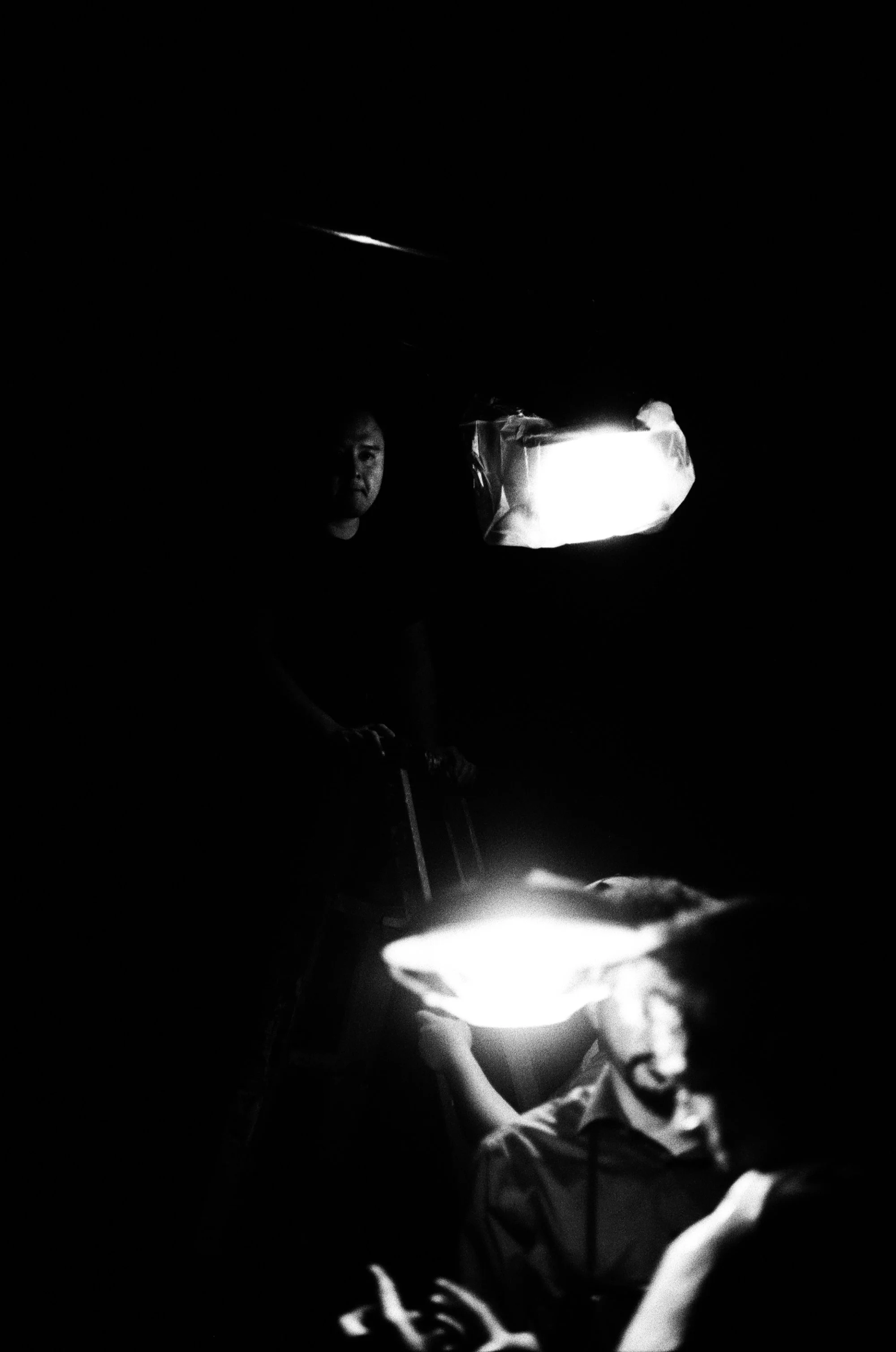 A dark room with three people dimly lit by glowing rectangular lamps, two are sitting and one is standing, with the person standing looking directly at the camera.
