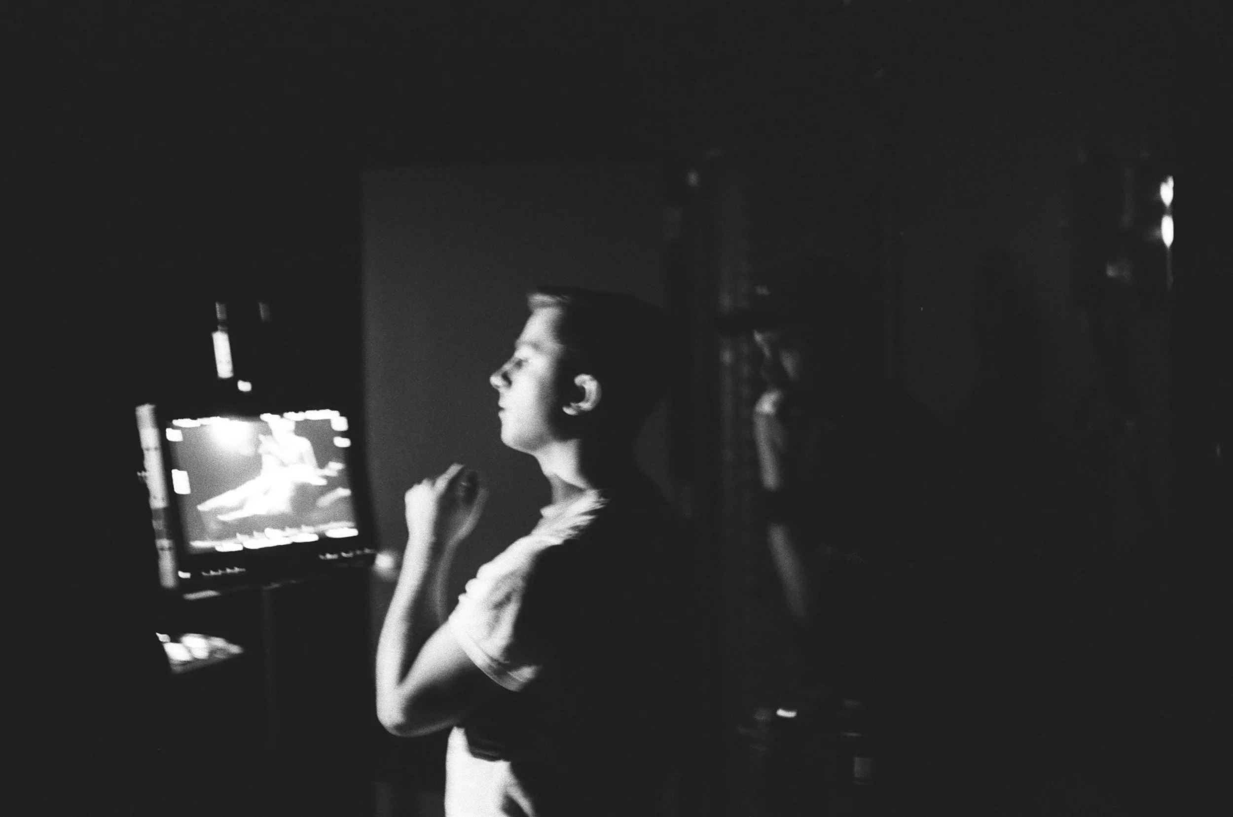 A person sitting in front of a monitor displaying a ballet dancer, captured in black and white, with soft lighting focusing on the person.