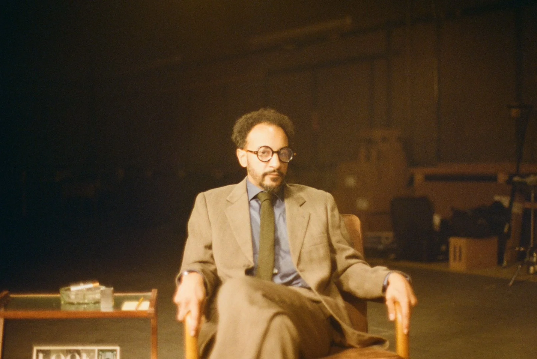 A man with glasses, curly hair, and a beard is sitting in a chair on a stage, wearing a brown suit, blue shirt, and dark tie, with a serious expression.