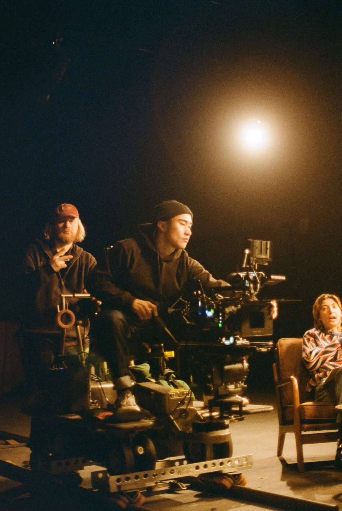 Three people on a film set: a man in a black hoodie and beanie operating a large camera, a man in a black hoodie and red cap making a peace sign, and a woman with short hair and a patterned shirt sitting on a chair, with a bright light overhead.