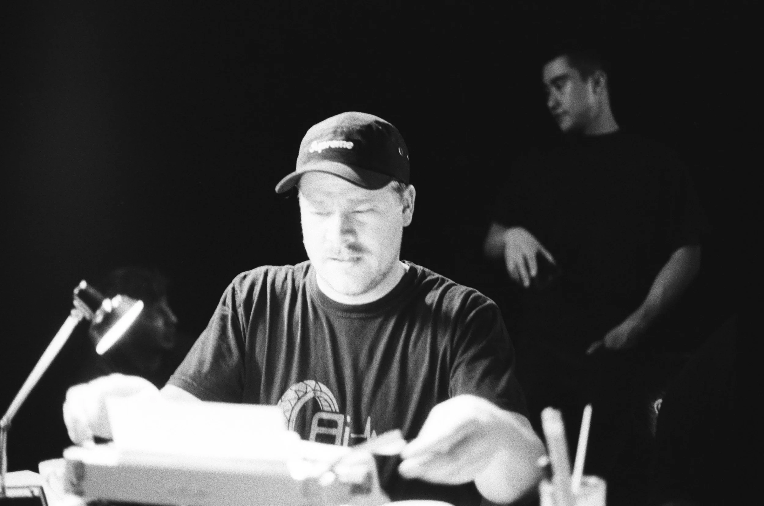 Black and white photo of three men in a dimly lit room. One man in the foreground wears a baseball cap and is working with electronic equipment, focusing on the task. Two other men are in the background, one sitting at a desk with a lamp, and another