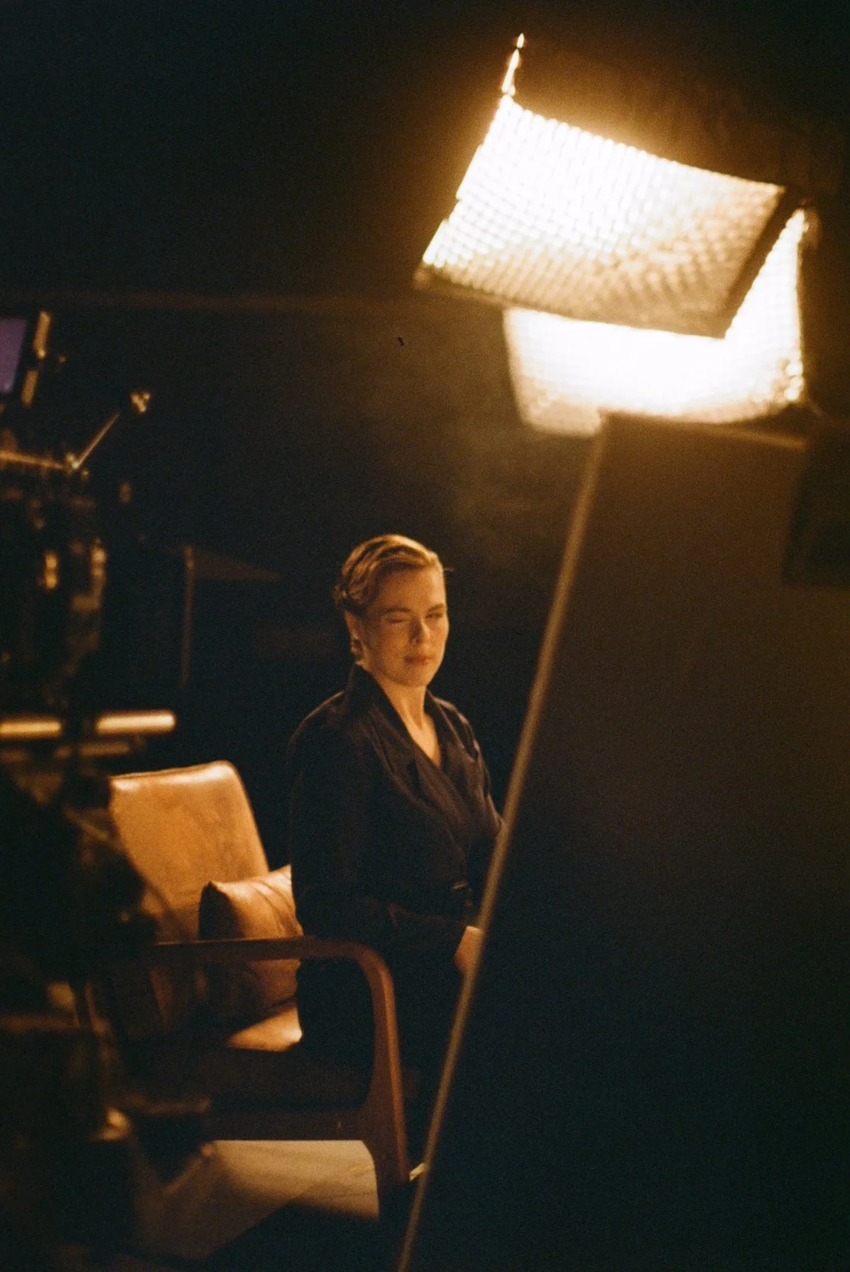 A woman sits in a leather chair in a dimly lit studio, illuminated by a large professional light. She is looking slightly towards the camera with a subtle smile.