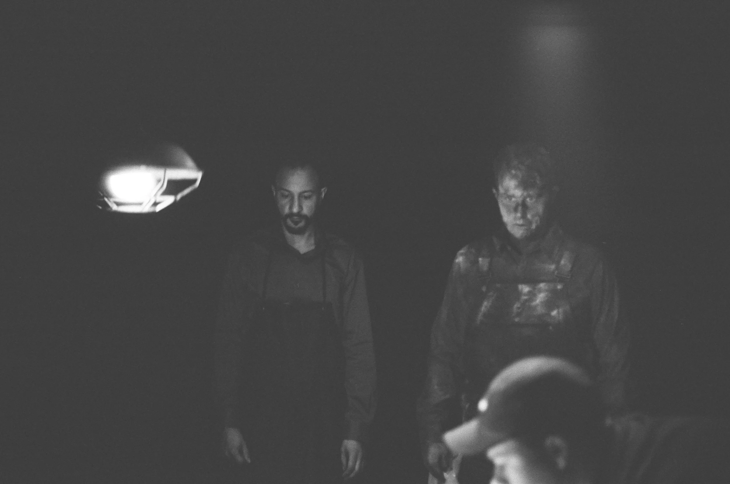Two men with dark clothing and serious expressions are standing in a dark space, illuminated by a small light source.