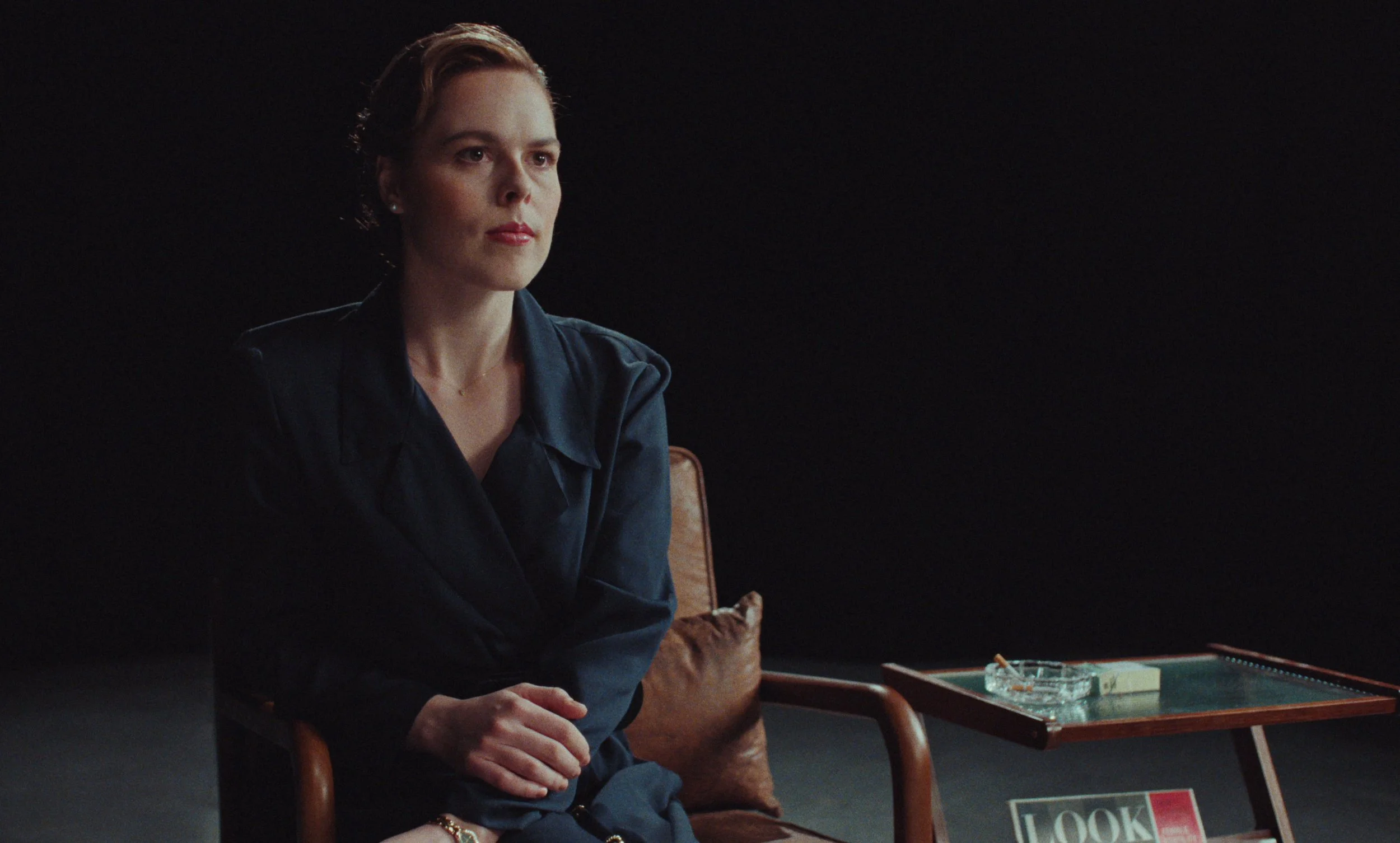 A woman with short red hair and a serious expression sitting in a dimly lit room wearing a dark blouse. She's seated on a wooden chair with a brown cushion. Next to her is a small wooden table with a glass ashtray, cigarette pack, and an ash, with a magazine titled "LOOK" visible underneath.