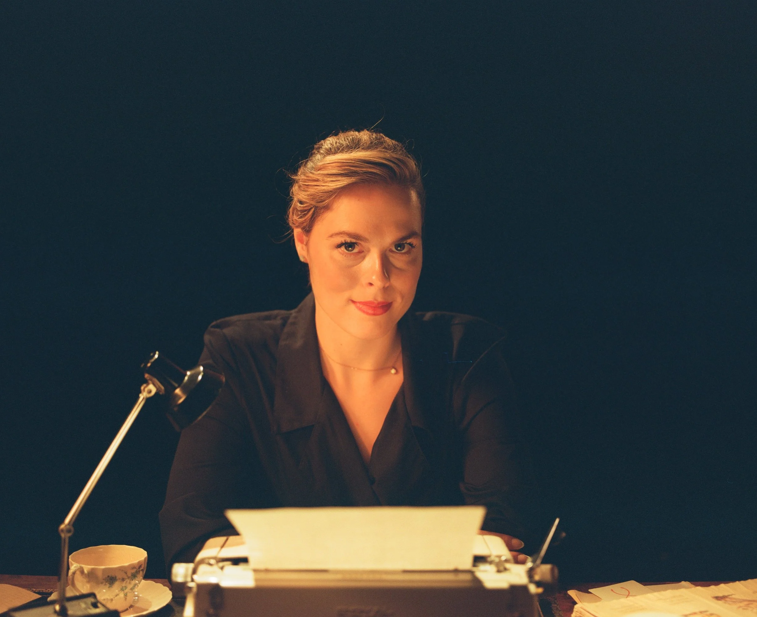 A woman with blonde hair styled in an updo, wearing a black blouse, sitting at a desk with a typewriter, a teacup, and papers, looking confidently at the camera against a dark background.