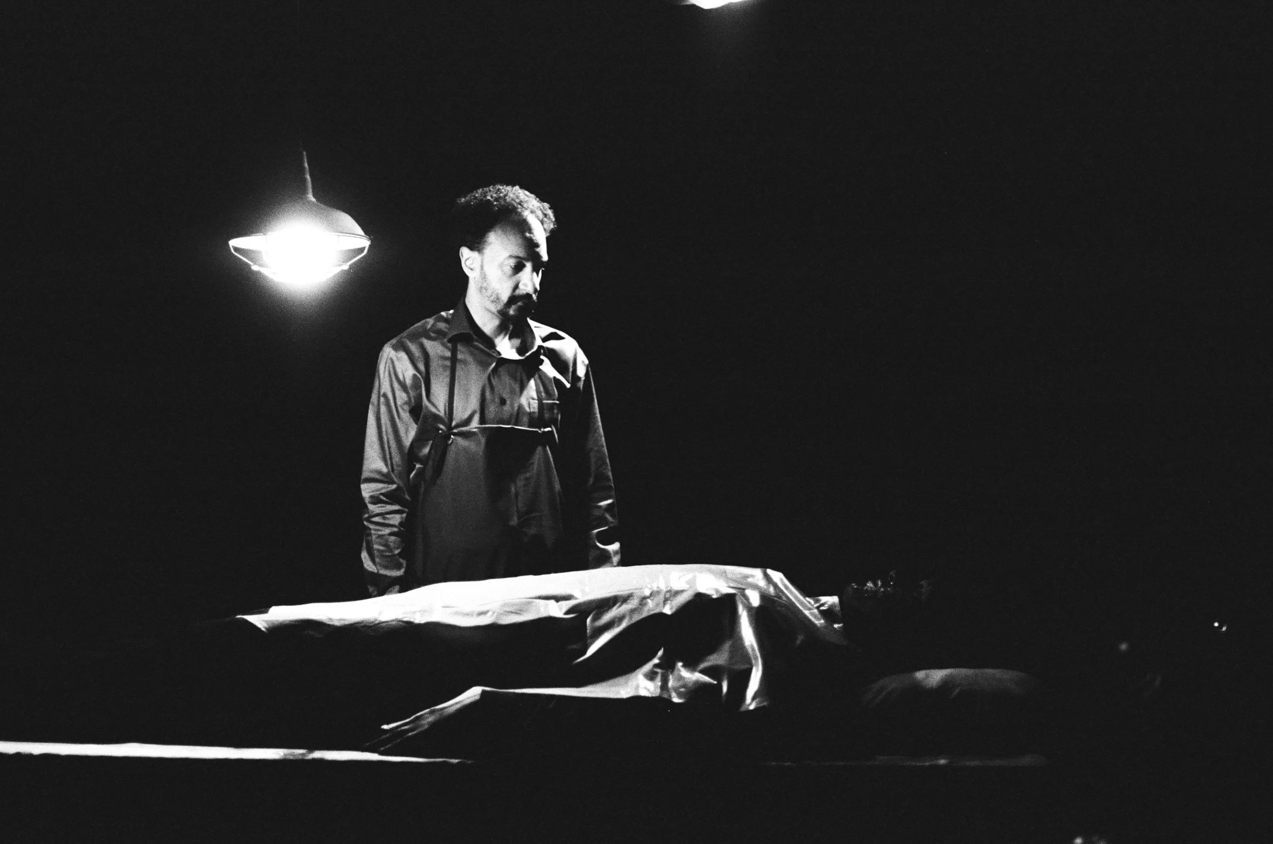 A man in a dark shirt looks down at a body covered with a sheet, lying on a table. The scene is dimly lit with a single bright overhead light.