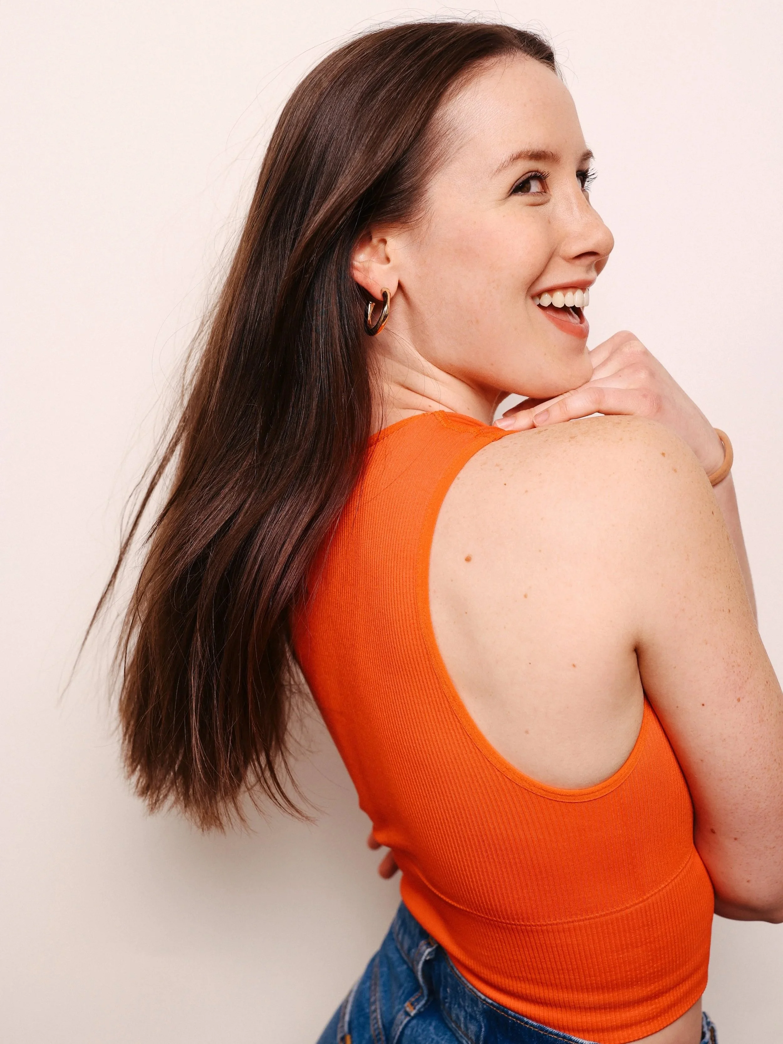A woman with long brown hair, wearing an orange sleeveless top and blue jeans, smiling and touching her chin, against a plain white background.