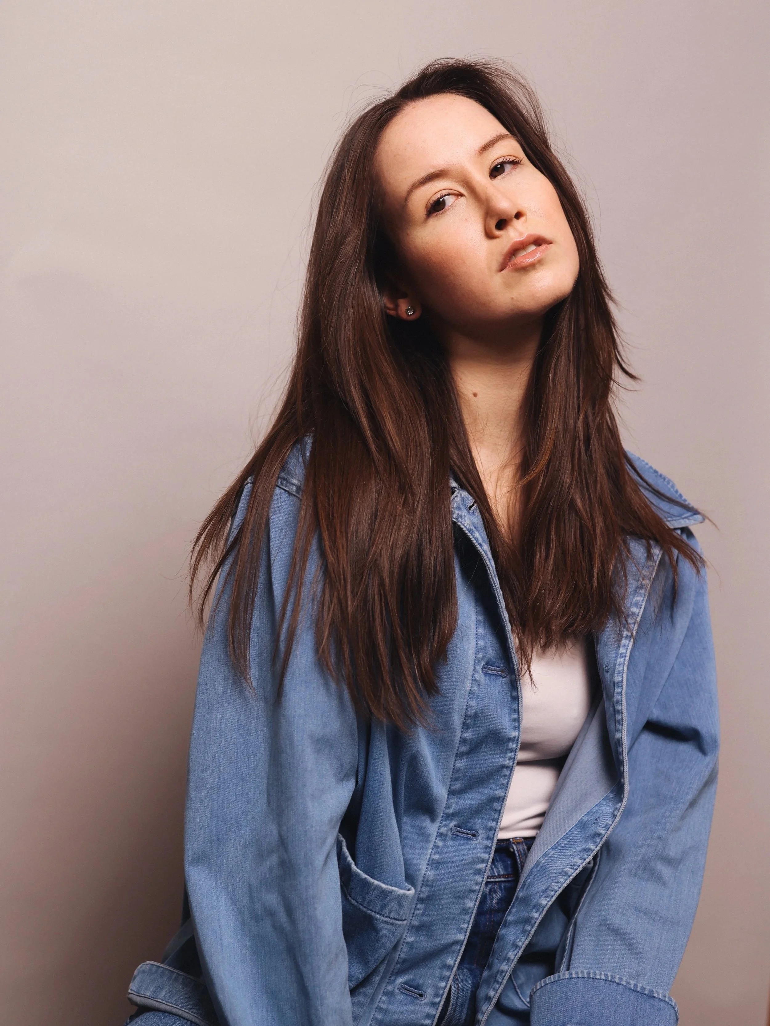 A young woman with long brown hair wearing a denim jacket and jeans, seated against a plain wall, looking slightly to the side.